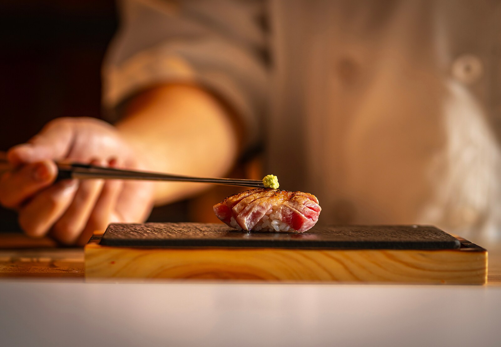 A pale, blurry chef’s hand holding chopsticks places a bead of wasabi atop a single peace of red-and-white sushi.