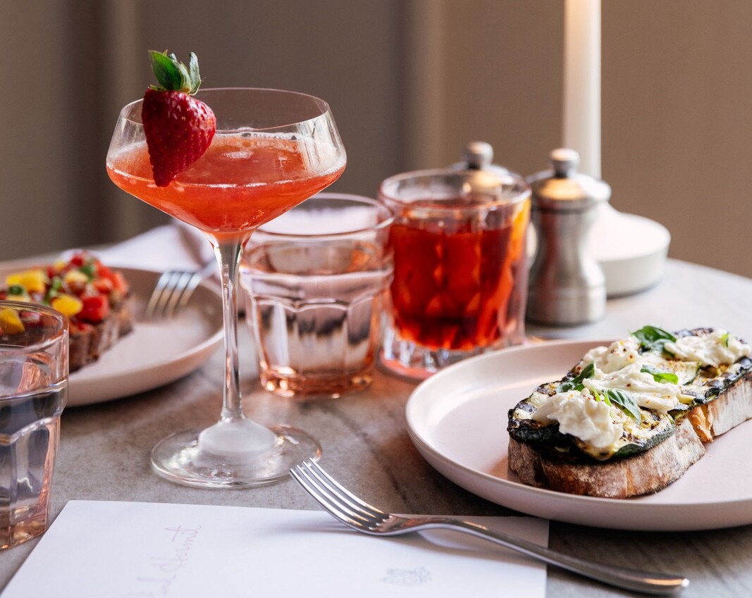 A table at The Pink Hermit is set with two red cocktails, one in a low glass and one in a stemmed glass, and plates of tartines.