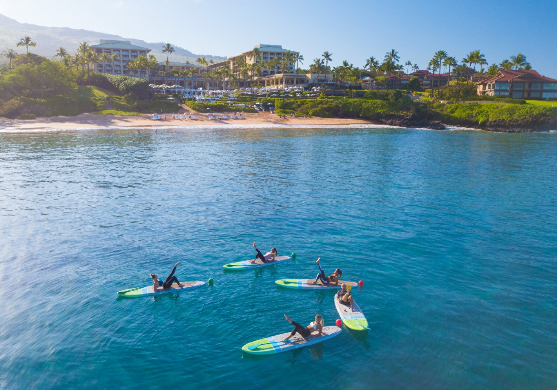 Aerial view of five women lying on paddleboards a few hundred feet offshore, lifting their feet in a yoga pose.
