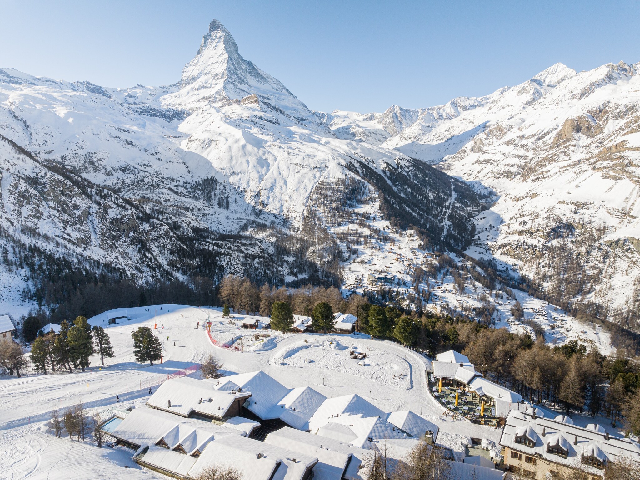 Aerial view of snow-covered buildings in a wooded valley at the base of ski slopes and steep mountains, including the Matterhorn.