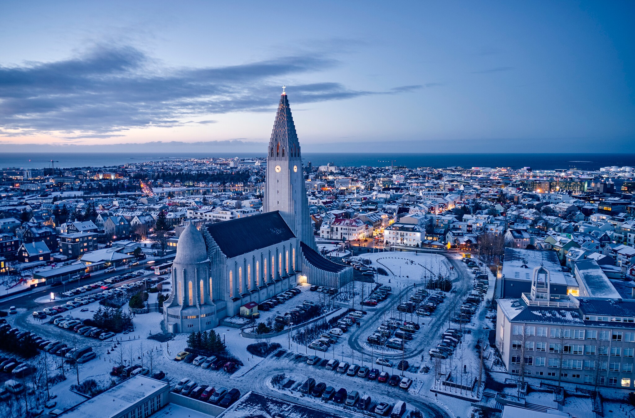 Aerial view over the snowy city and especially Hallgrímskirkja, a tall church with a spire and wings tapering to the side like a pipe organ.