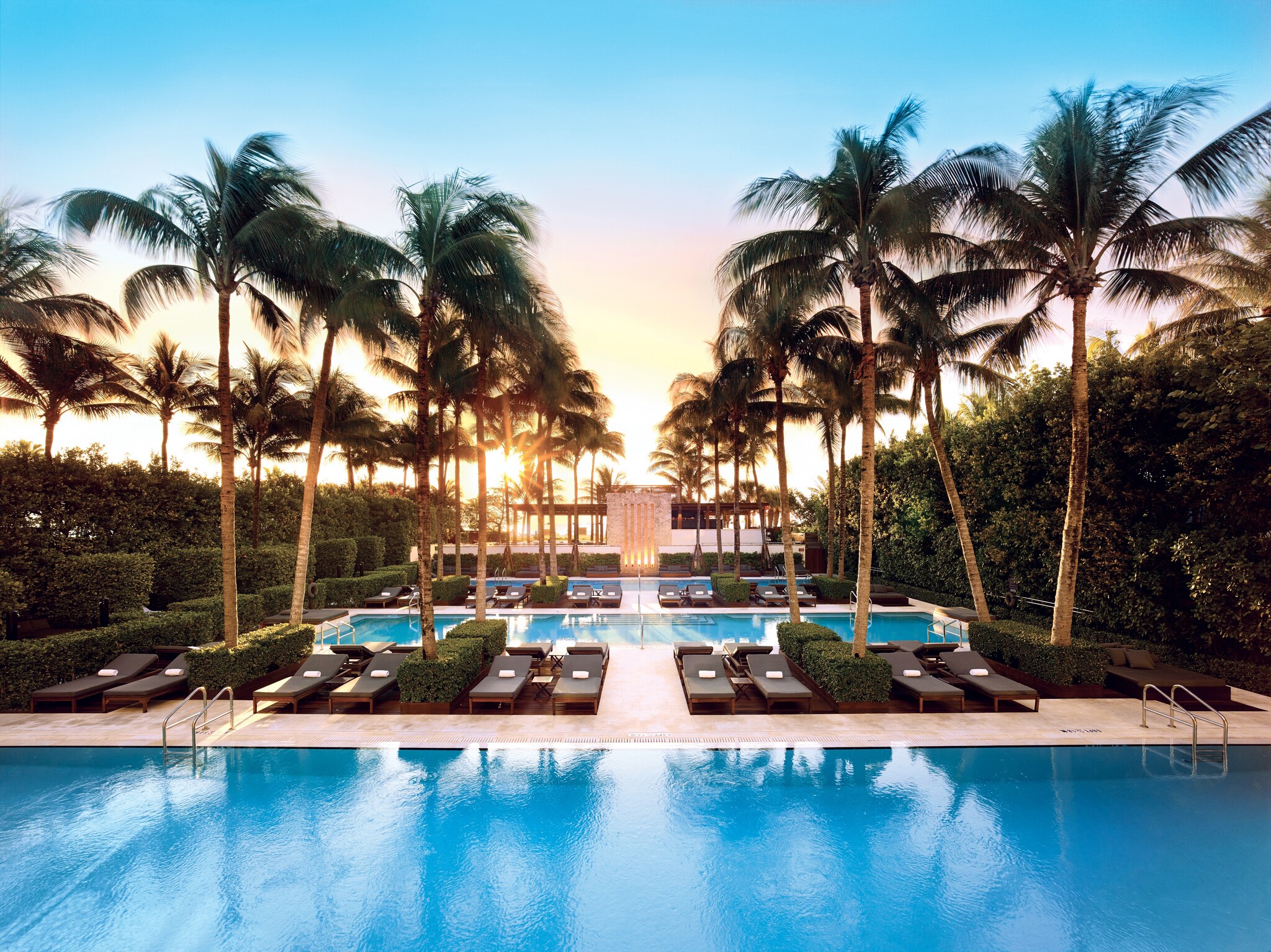 At sunrise, three medium-sized pools in a row stretch into the distance, with palm trees, sunbeds, and shrubs in between each.