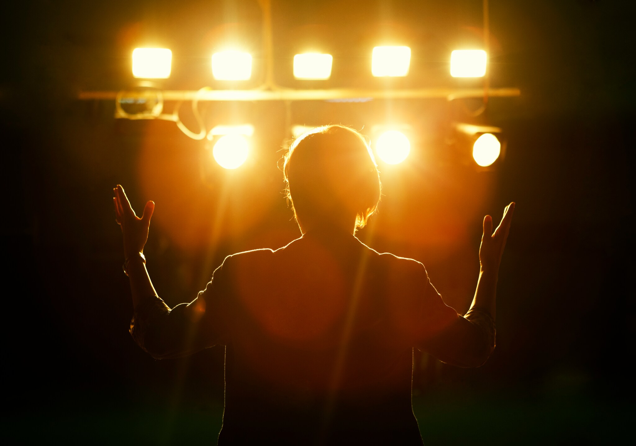 View from behind of a thin performer with short hair holding their arms up to the sides, silhouetted in front of a bank of stage lights.