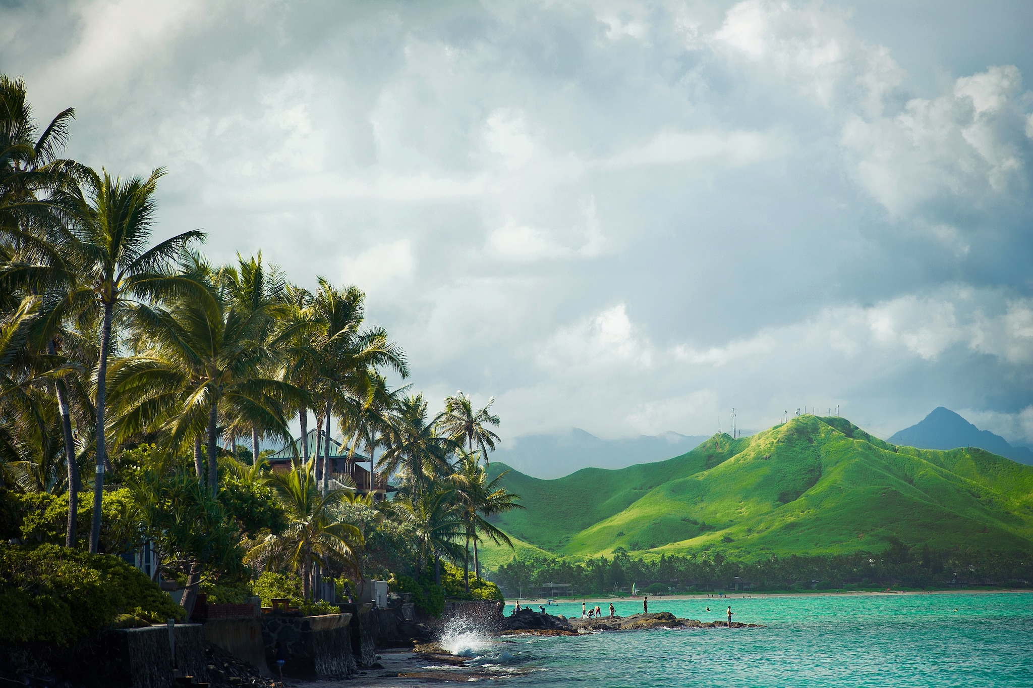 On a palm-fringed beach, visitors climb on volcanic rock. Beyond, a bright green mountain rises into a cloudy sky.