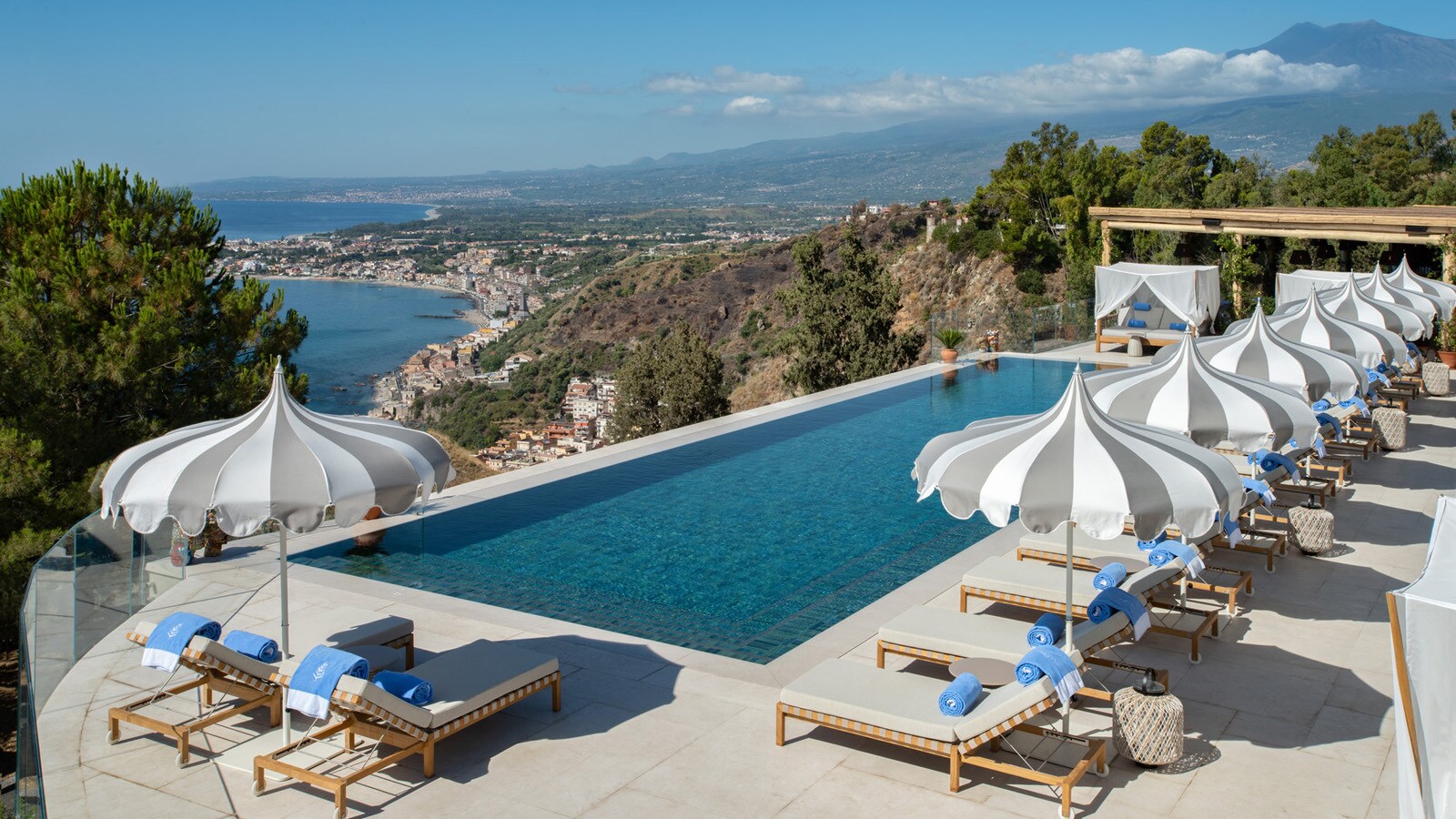 A cliffside infinity pool is surrounded by gray and white umbrellas and white sun beds covered in blue towels.