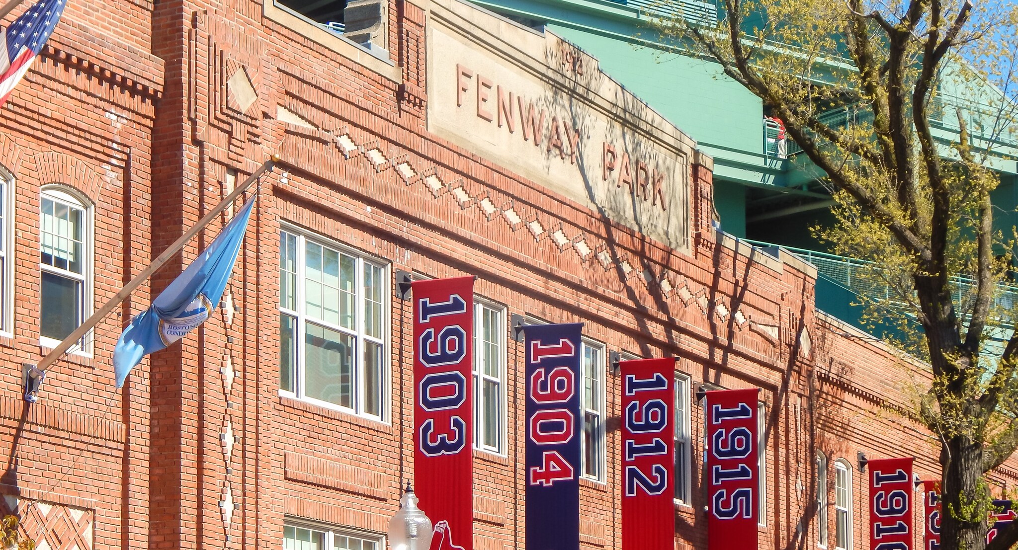The facade of Fenway Park is red brick with tan accents. Flags along the side list the years of Red Sox AL pennants in red, white, and blue.
