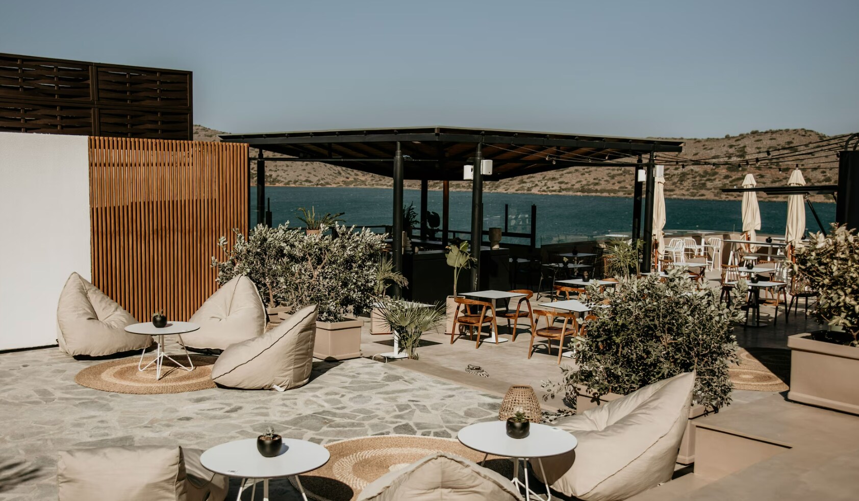 The Amphiteatro at Domes Aulus Elounda is an outdoor area with clusters of beige beanbags around round white tables and woven rugs.
