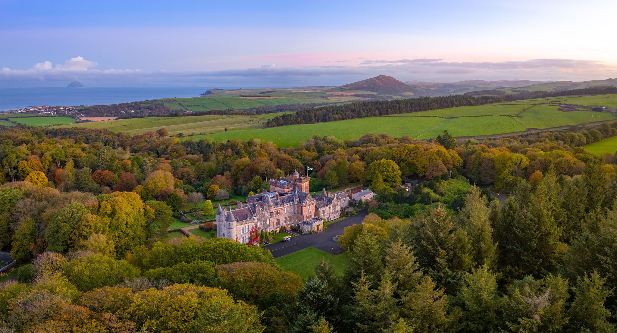 Aerial view of Truestone Glenapp Castle Limited, a castle nestled in a forest with the water of the Firth of Clyde visible in the distance.