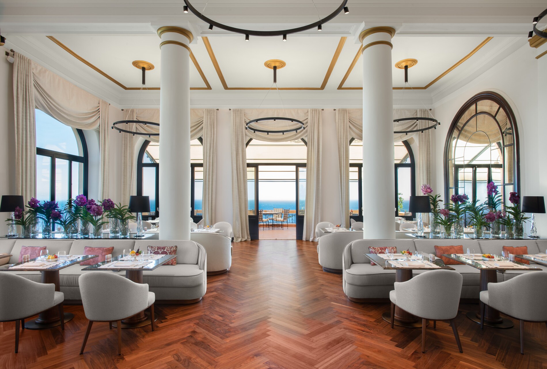 The Principe Cerami dining room has parquet floors, white columns, and light gray chairs and couches arranged around square tables.