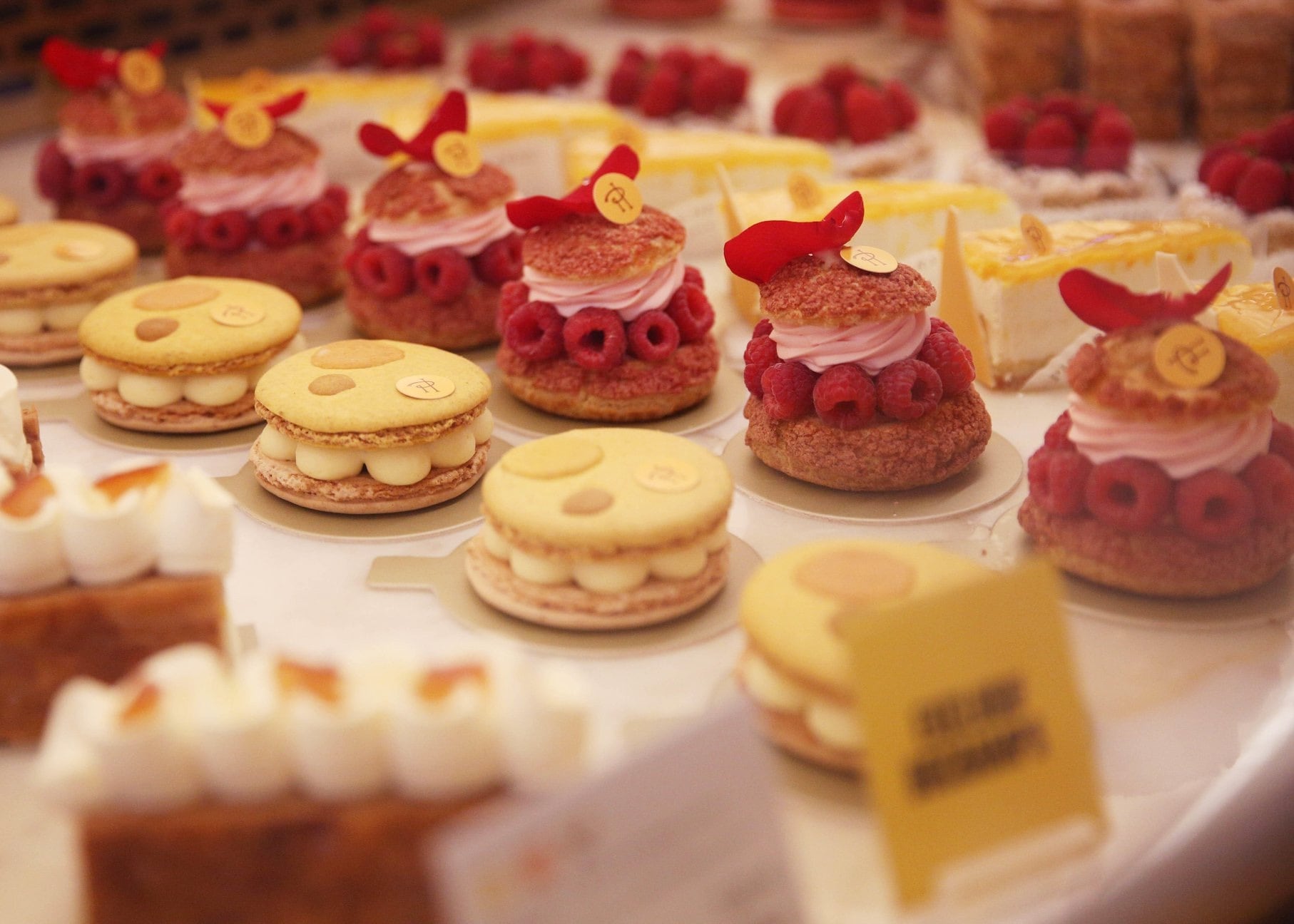 Rows of yellow macarons and pink pastries with cakes, berries, and frosting are arrayed on paper circles under warm light.