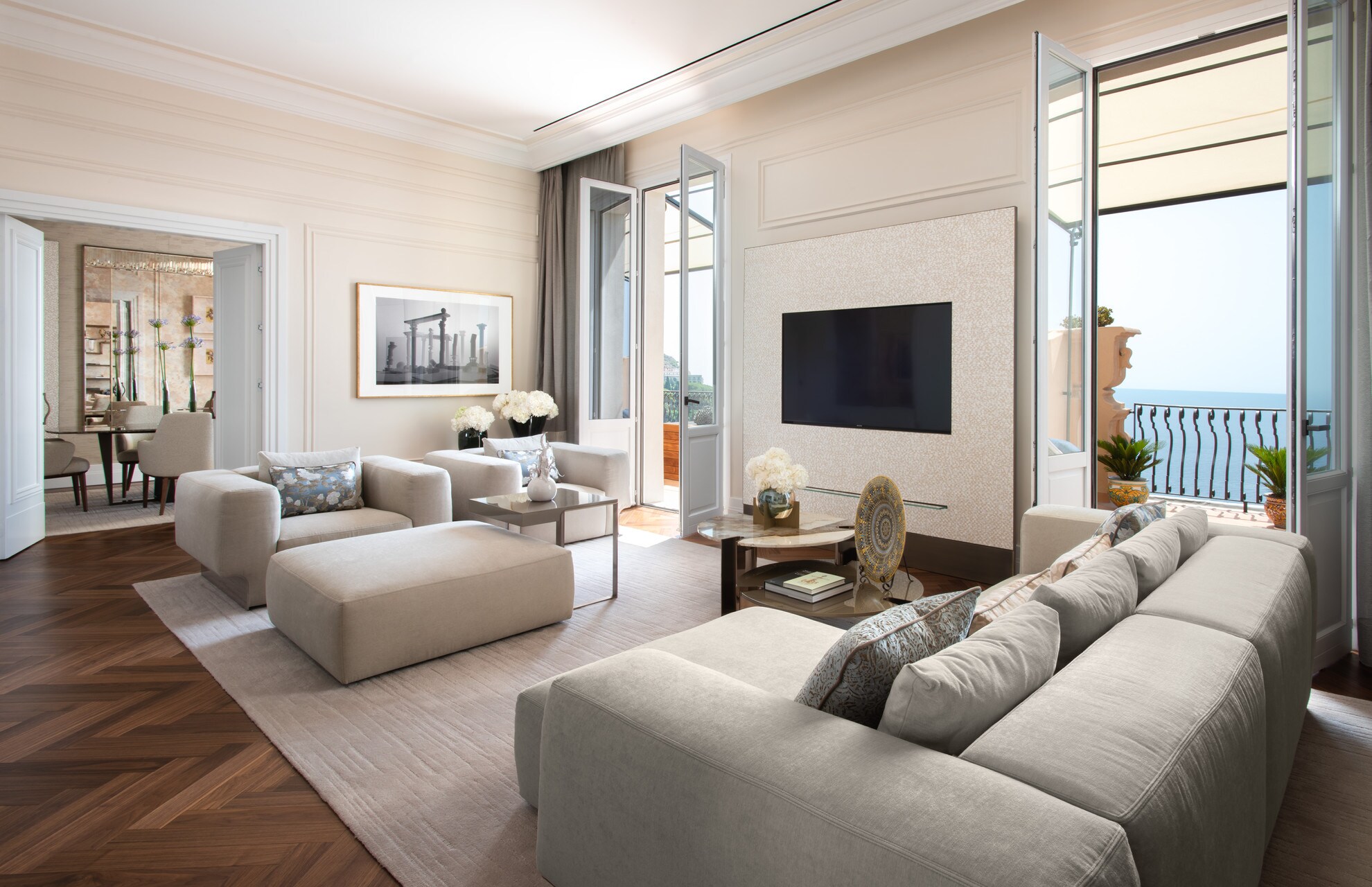 The Royal Suite Living Area has boxy light-gray couches arranged around a wall-mounted television. Tall glass doors open onto a balcony.