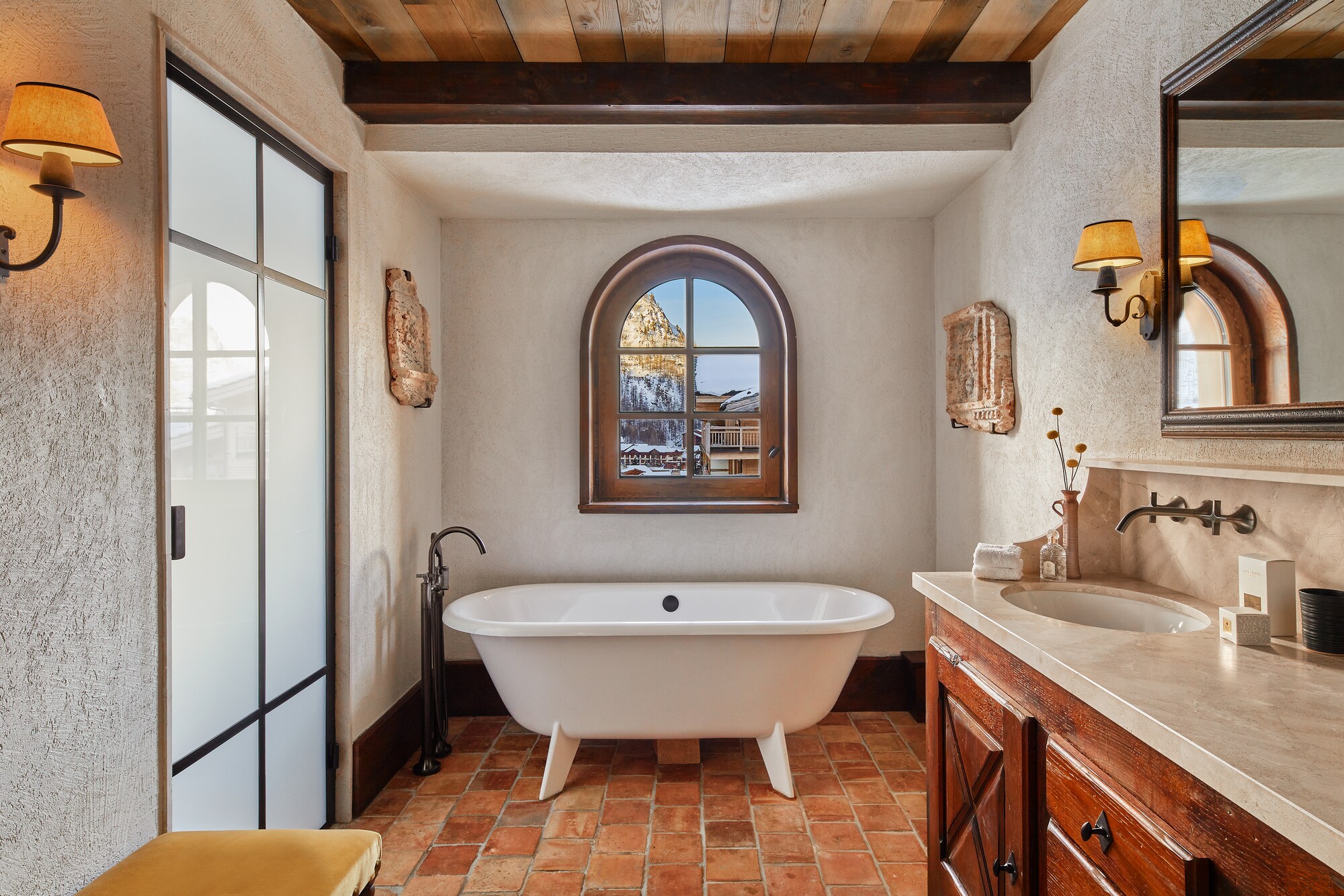 At one end of a bathroom with textured walls, brick floors, and marble countertops, a freestanding tub sits under a window.