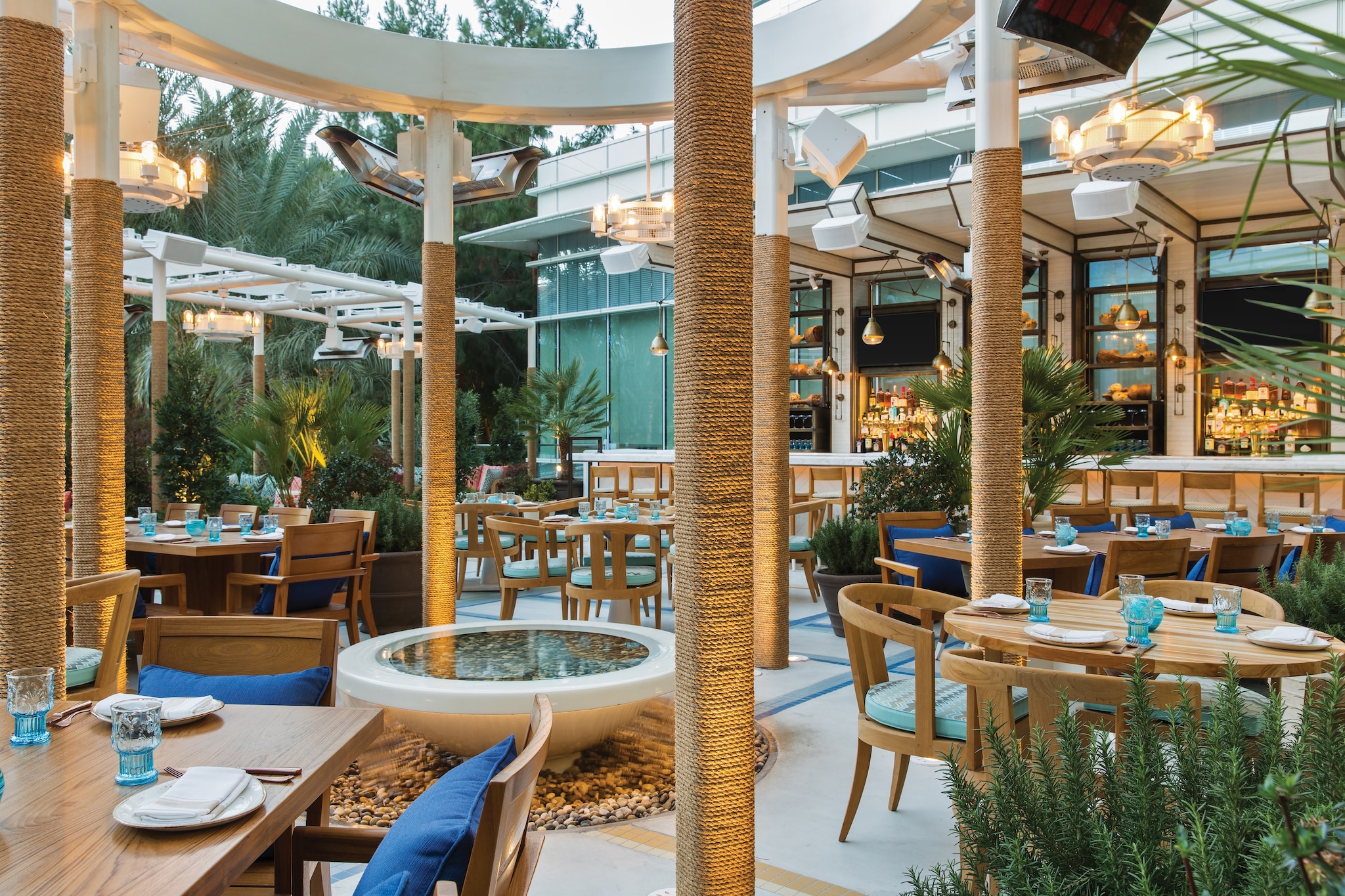 Salt & Ivy Café and Patio Bar is filled with potted plants and small wooden tables and chairs under pergolas with wicker-wrapped pillars.