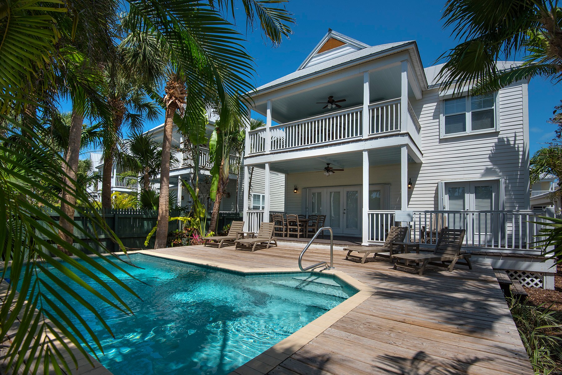Exterior view of the Sanctuary Village Villa, a two-story white building with a railed balcony and a pool shaded by palm trees.