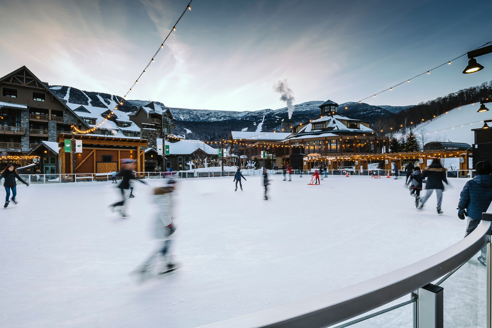 Blurred skaters cross an ice rink under lit strings of lights. Across the rink, lodges and mountains are covered in snow.