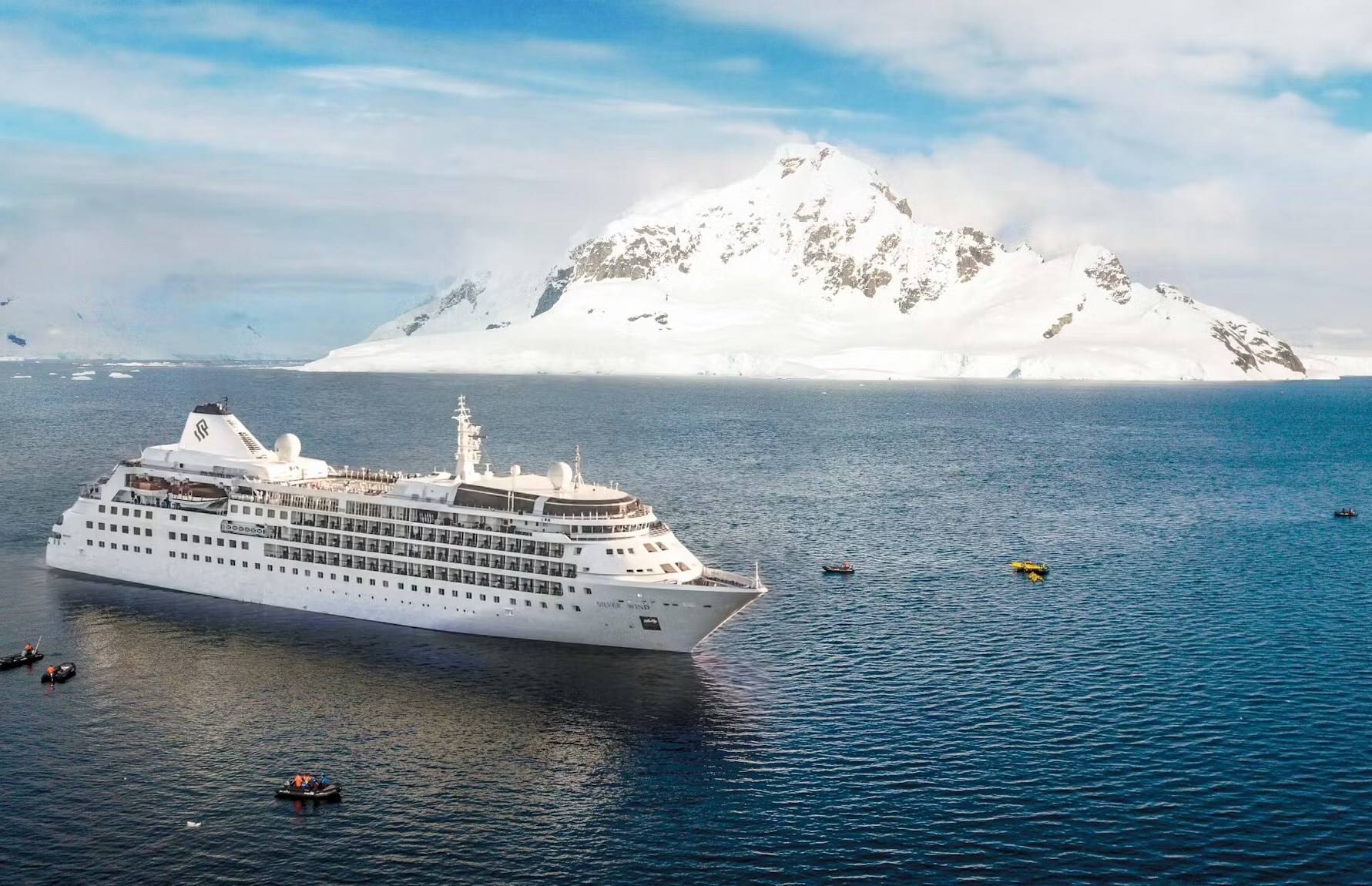 A white cruise ship in the water in front of a snowy mountain. Small inflatable boats paddle around the ship.