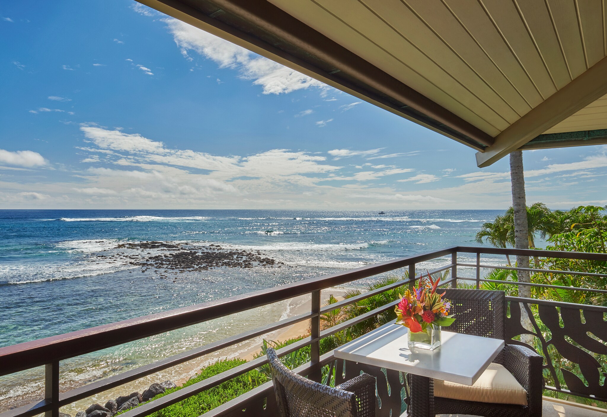 View from a guest room balcony of the beach. On the balcony, two wicker chairs are seated next to a small table with a vase of flowers.