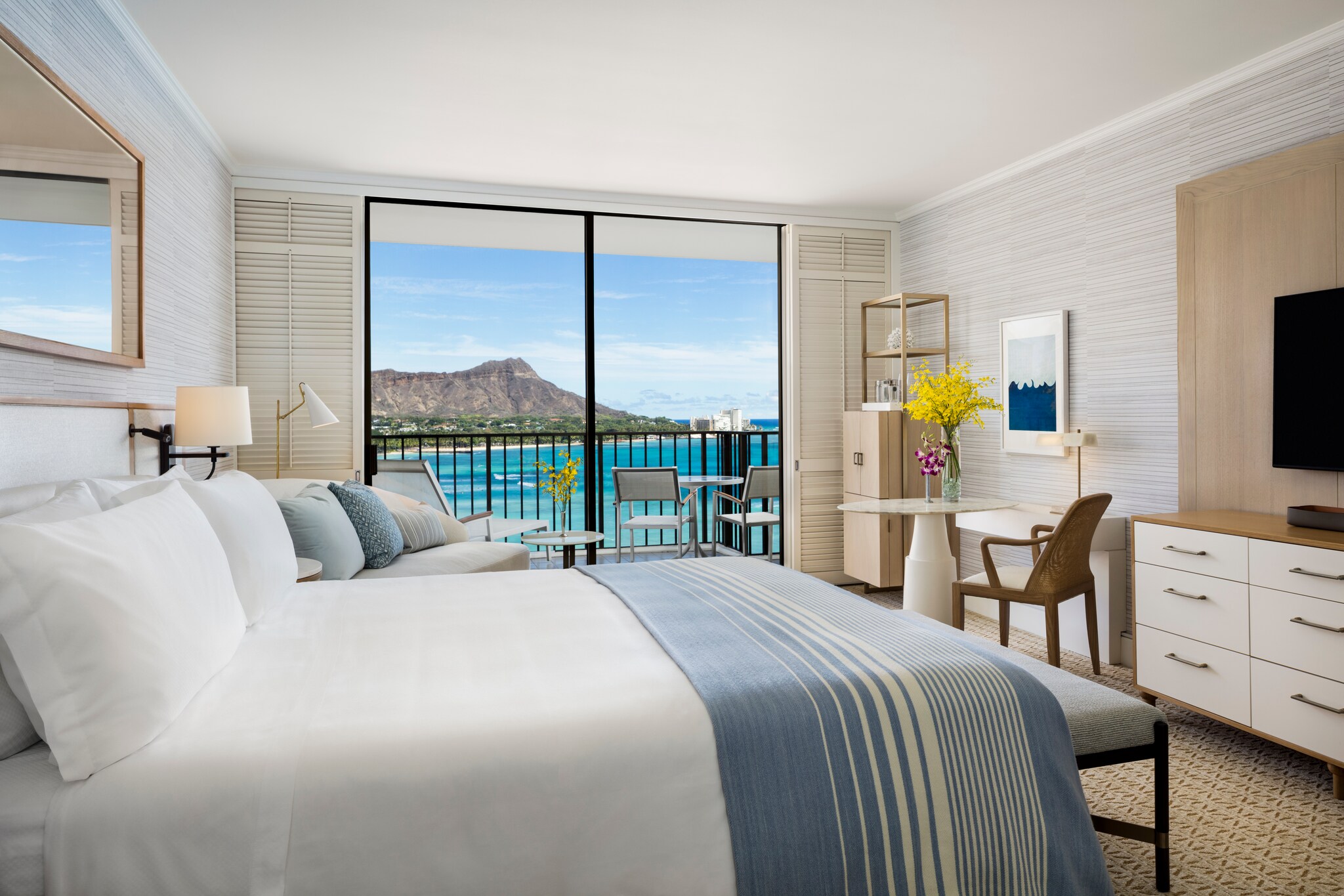 Out the window of a Diamond Head Ocean Guestroom, the namesake volcano is framed. Inside, the room is decorated white, blue, and brown.