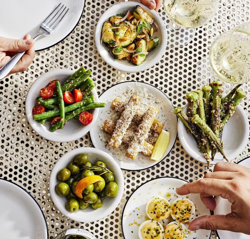 Overhead view of myriad dishes, mostly olives and vegetables lightly charred and crusted with seasoning. A few diners’ hands pick at the edges.