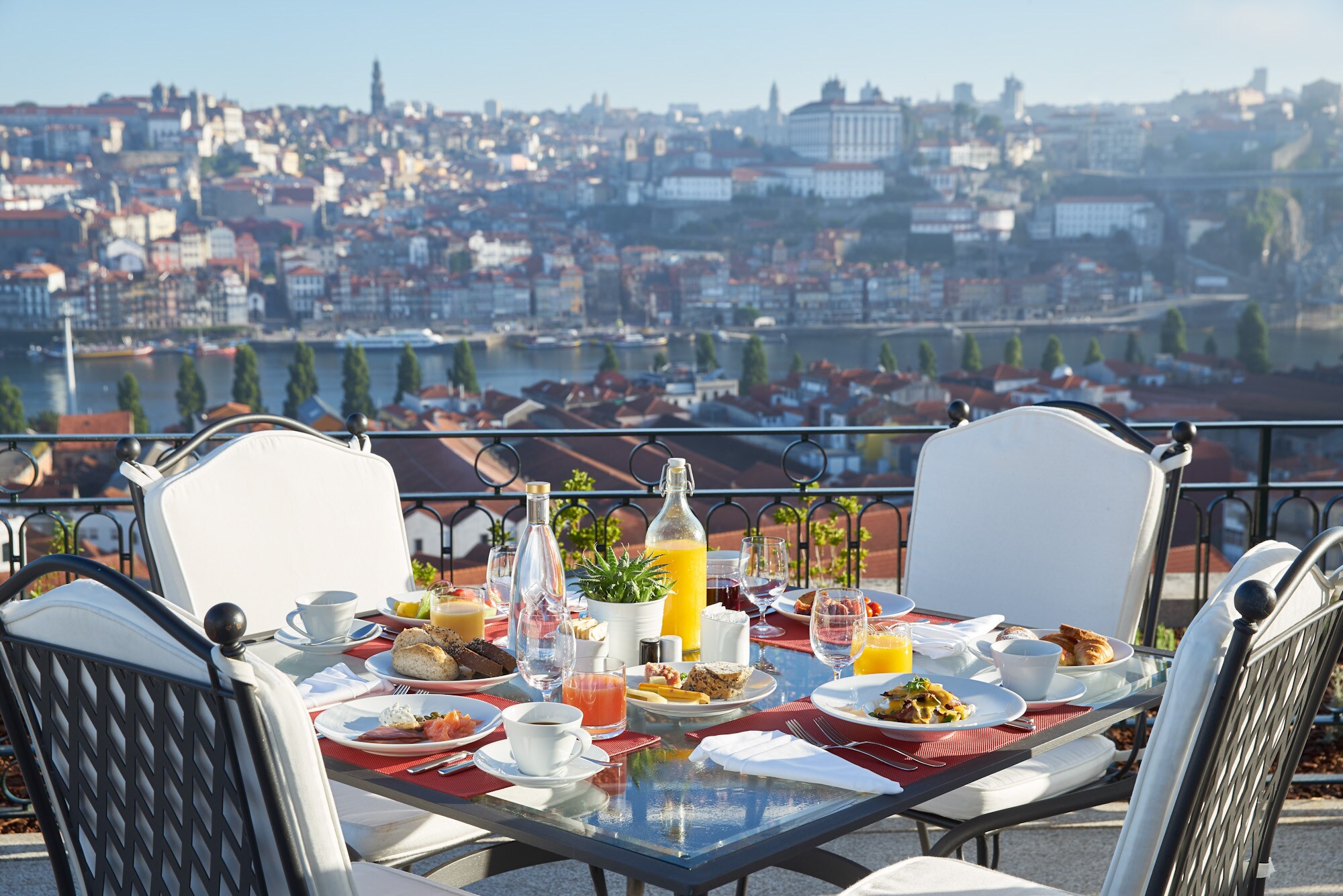 On a terrace overlooking the city on a sunny day, a square table is set with breakfast, including juice, pastries, and coffee.