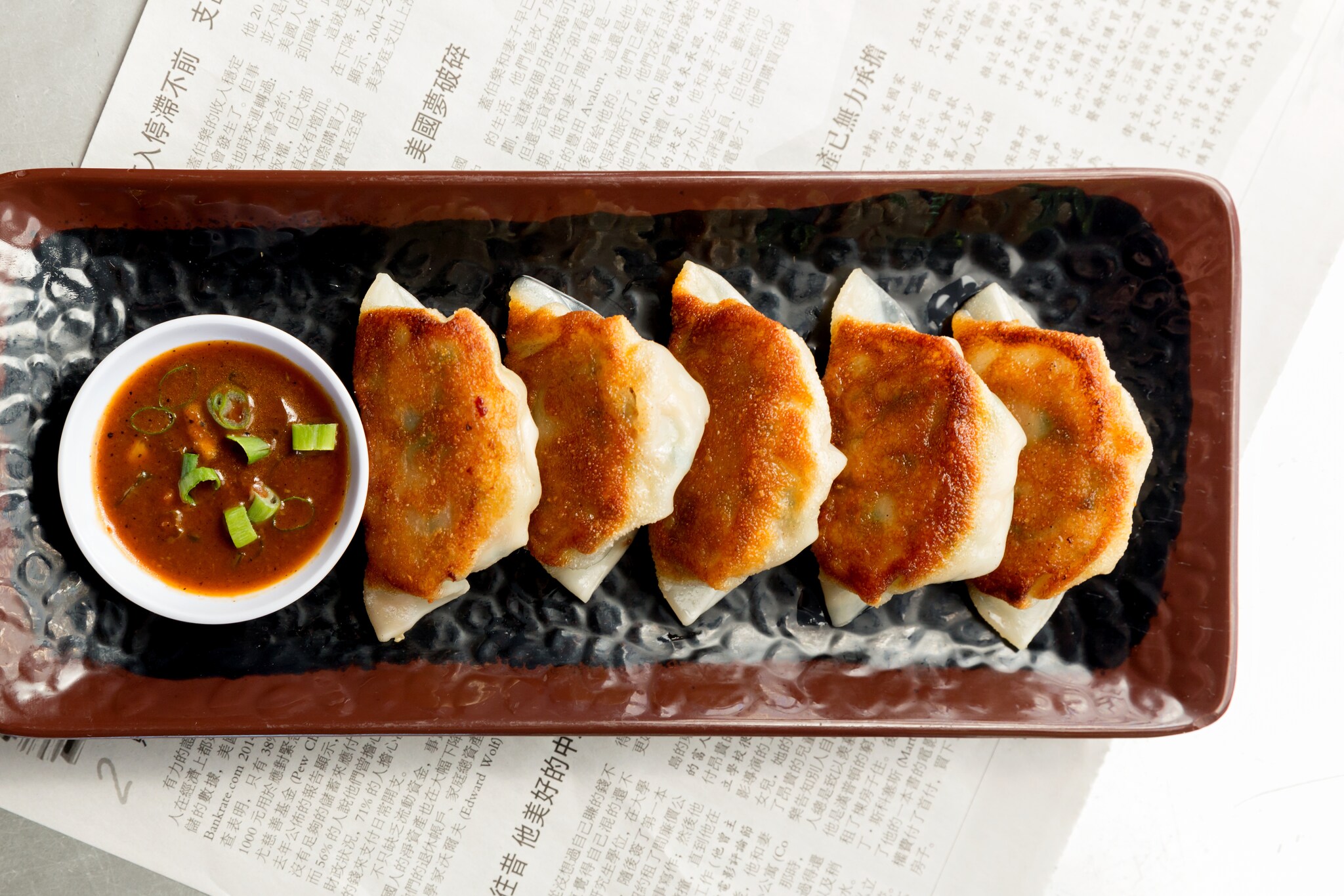 Overhead view of a black-and-brown rectangular platter of browned dumplings, served with a bowl of red-brown sauce with green onions.