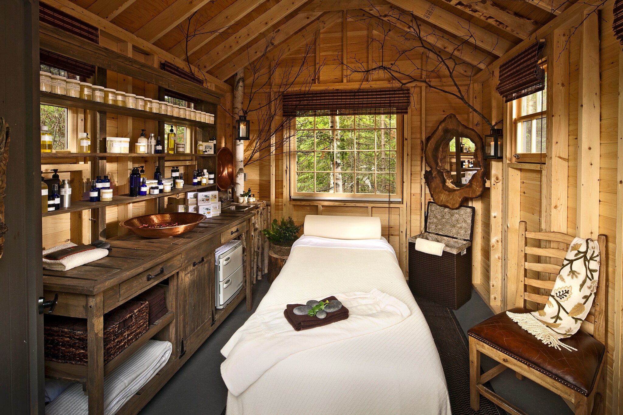 A spa room is built of knotted wood, ornamented with an apothecary shelf, black tree branches, and a log-rimmed mirror around a white bed.