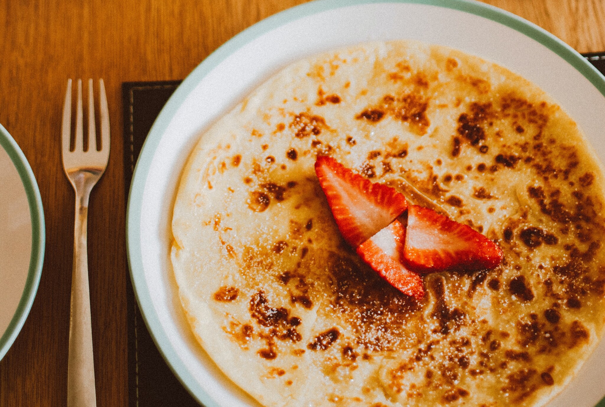 A crepe sprinkled with powdered sugar and with three wedges of strawberry rests on a white and green plate on a wooden table with a fork.