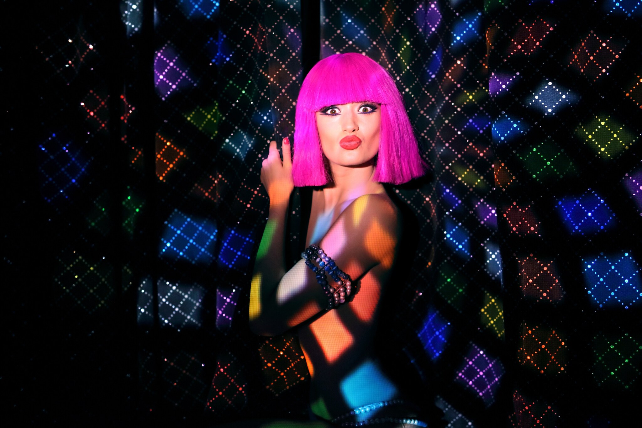 A dancer in a hot-pink bob wig and scant clothing clutches at curtains and makes a surprised kissy face at the camera, awash in colorful lights.