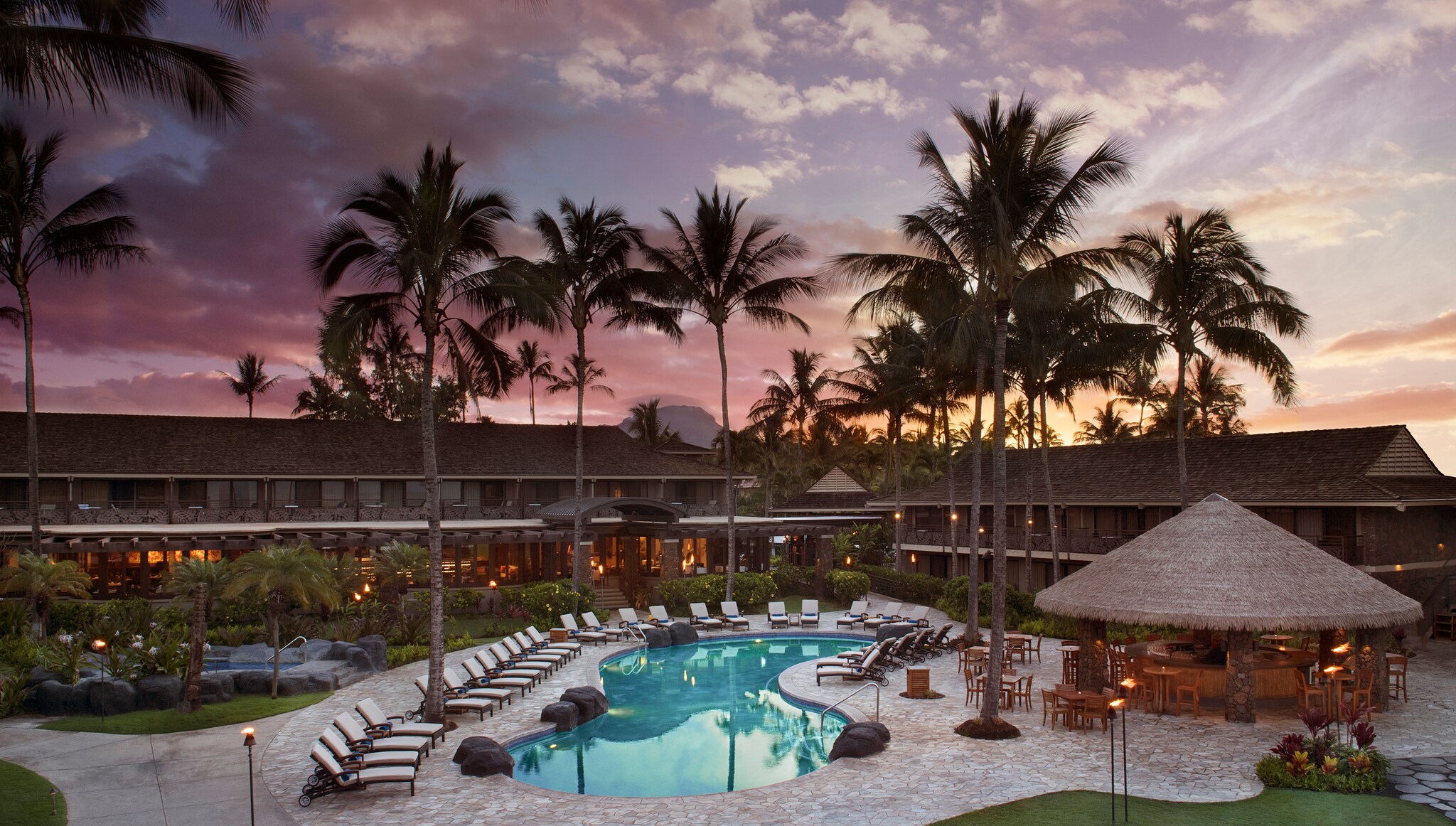 Aerial view of the hotel as the sun sets. Palm trees around the bean-shaped pool are silhouetted in front of the two-story hotel.