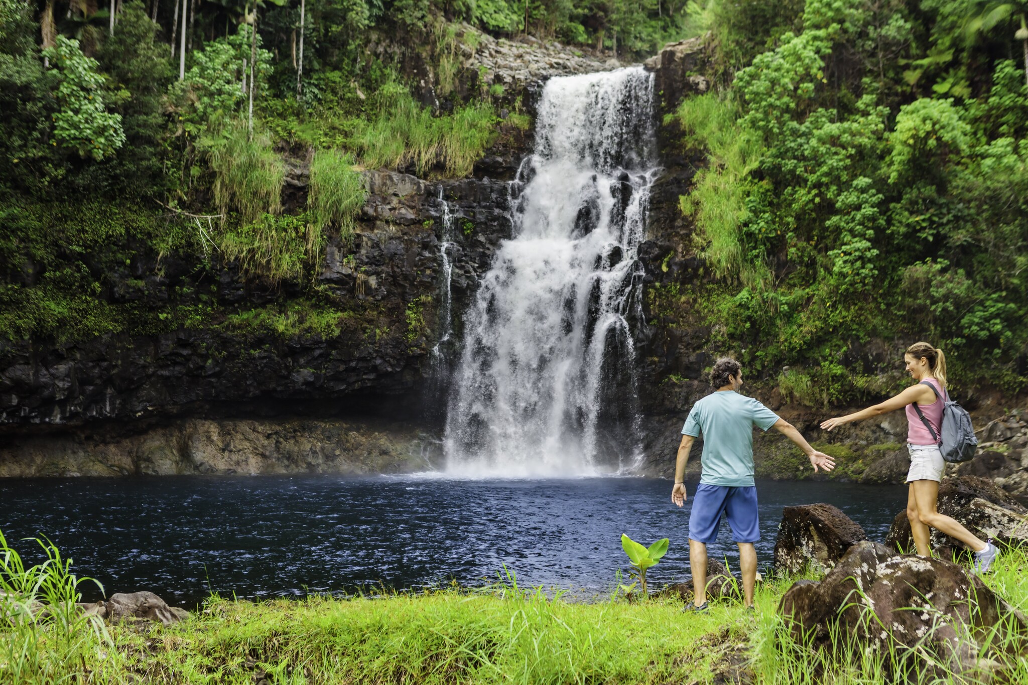 A white man and a white woman hold their hands out to each other as they walk around a pool in front of a waterfall.