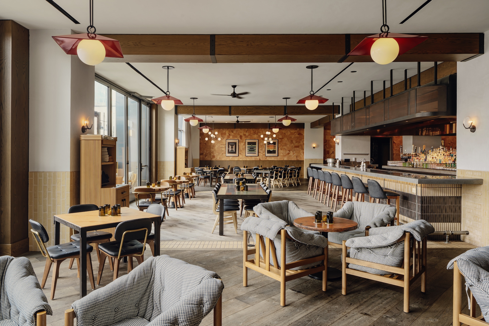 An expansive hotel restaurant features a warm neutral palette, a long tiled bar, and sunlight streaming in from floor-to-ceiling windows.