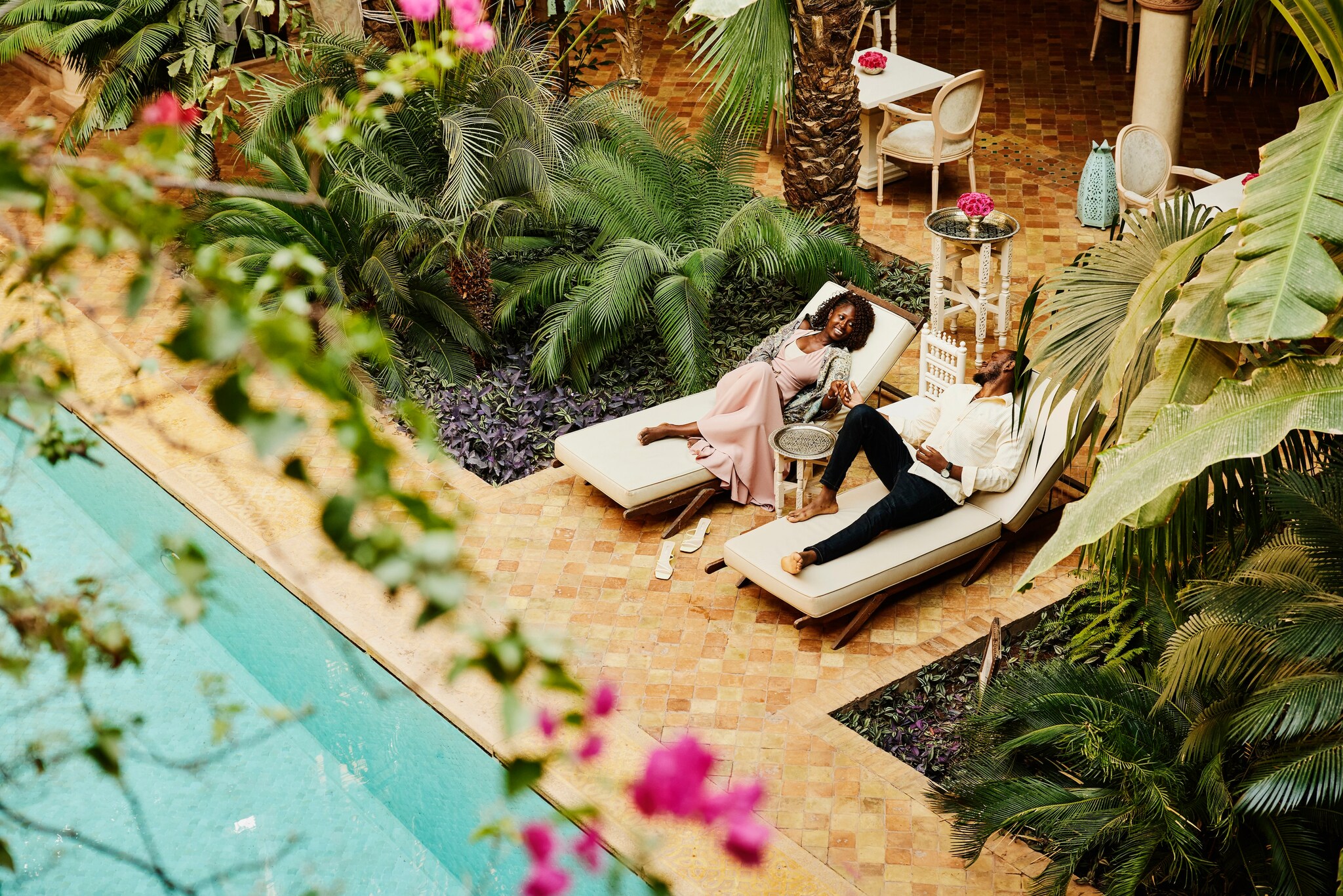 A Black couple holds hands from separate sun loungers next to a pool framed by lush tropical plants.