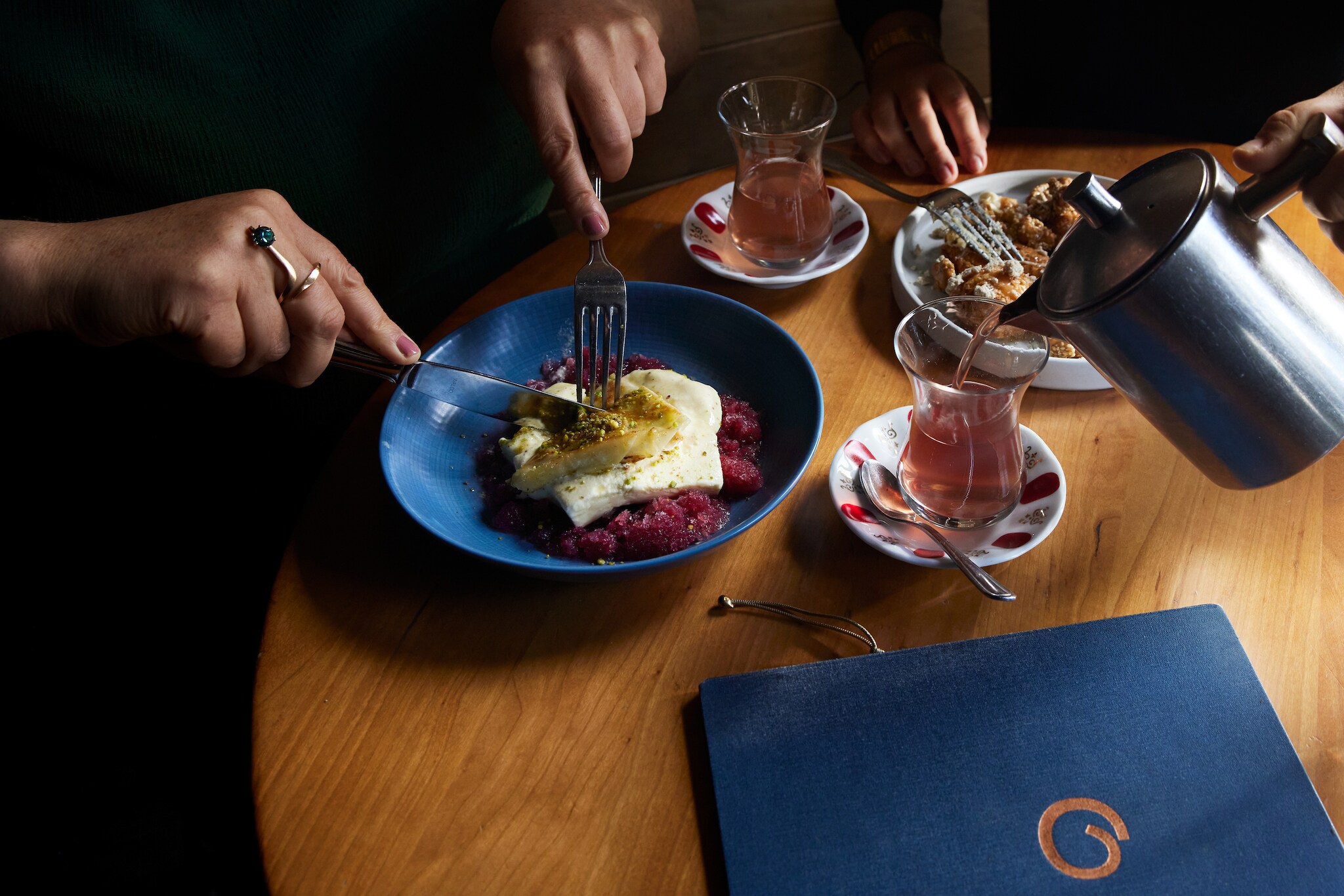 Tan hands use a fork and knife to cut into a pistachio katmer layered on top of white saffron ice cream and a bed of pomegranate granité.