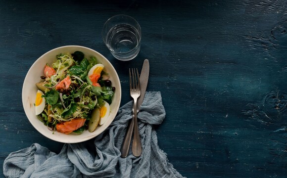 Overhead view of a salad in a white dish on a teal wooden table. The salad includes dark greens, wedges of hard-boiled egg, and salmon.