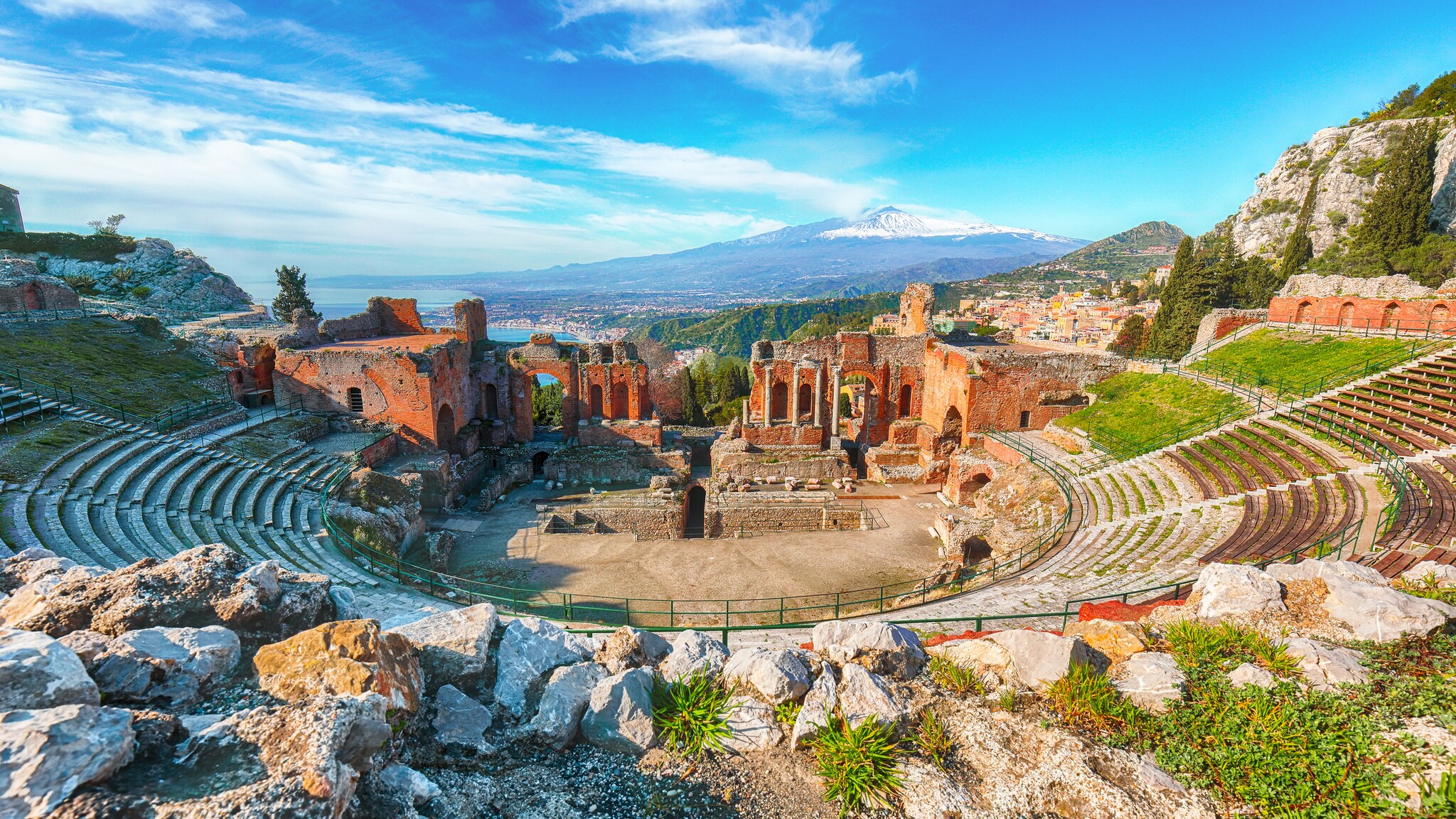 View from the upper stands of the ancient Greek theater of Taormina, a set of open-air ruins built of red and white stone.