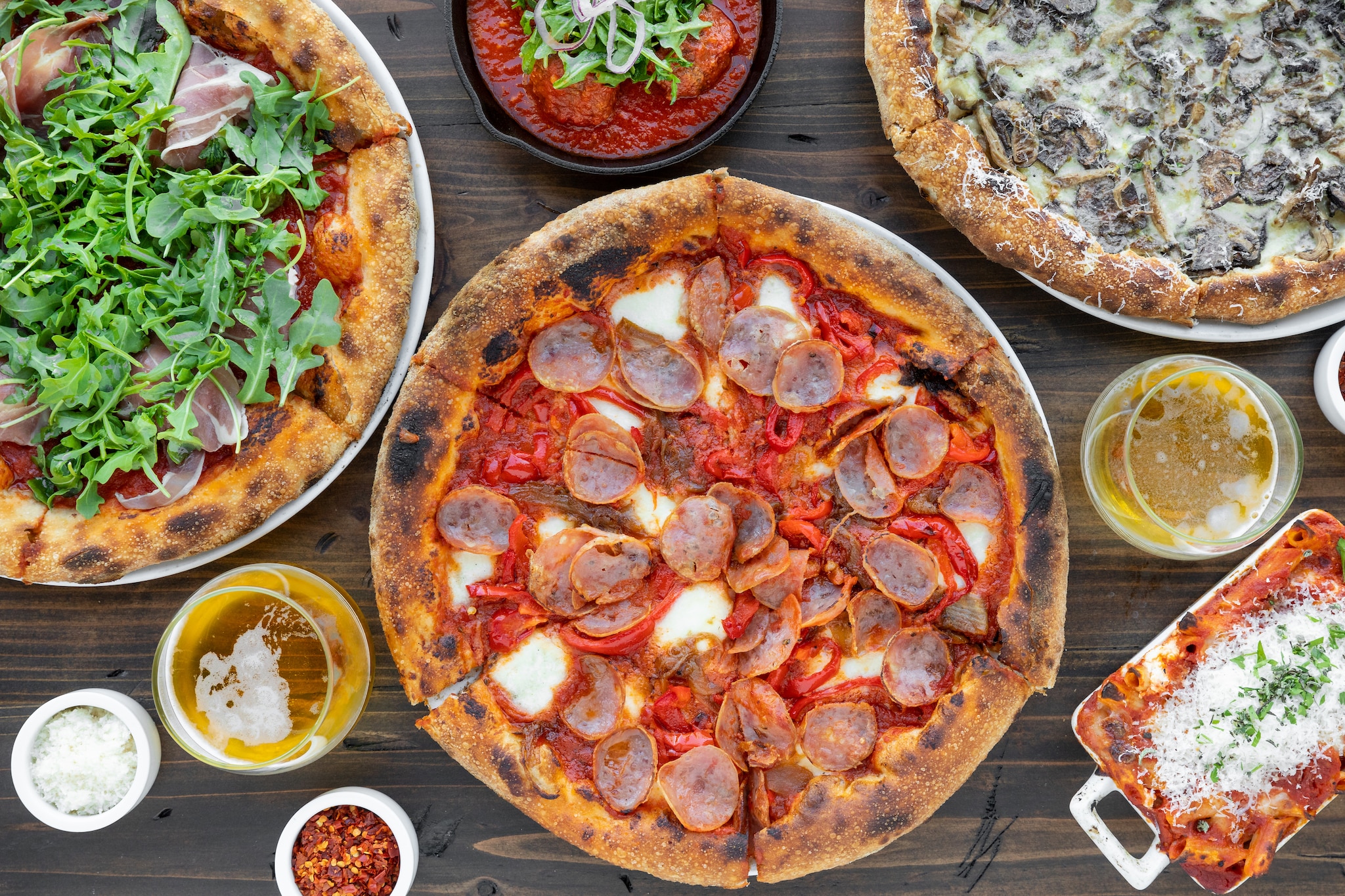 Overhead view of several pizzas, one with pepperoni, one with arugula, and one with mushrooms, accompanied by white wine and parmesan.