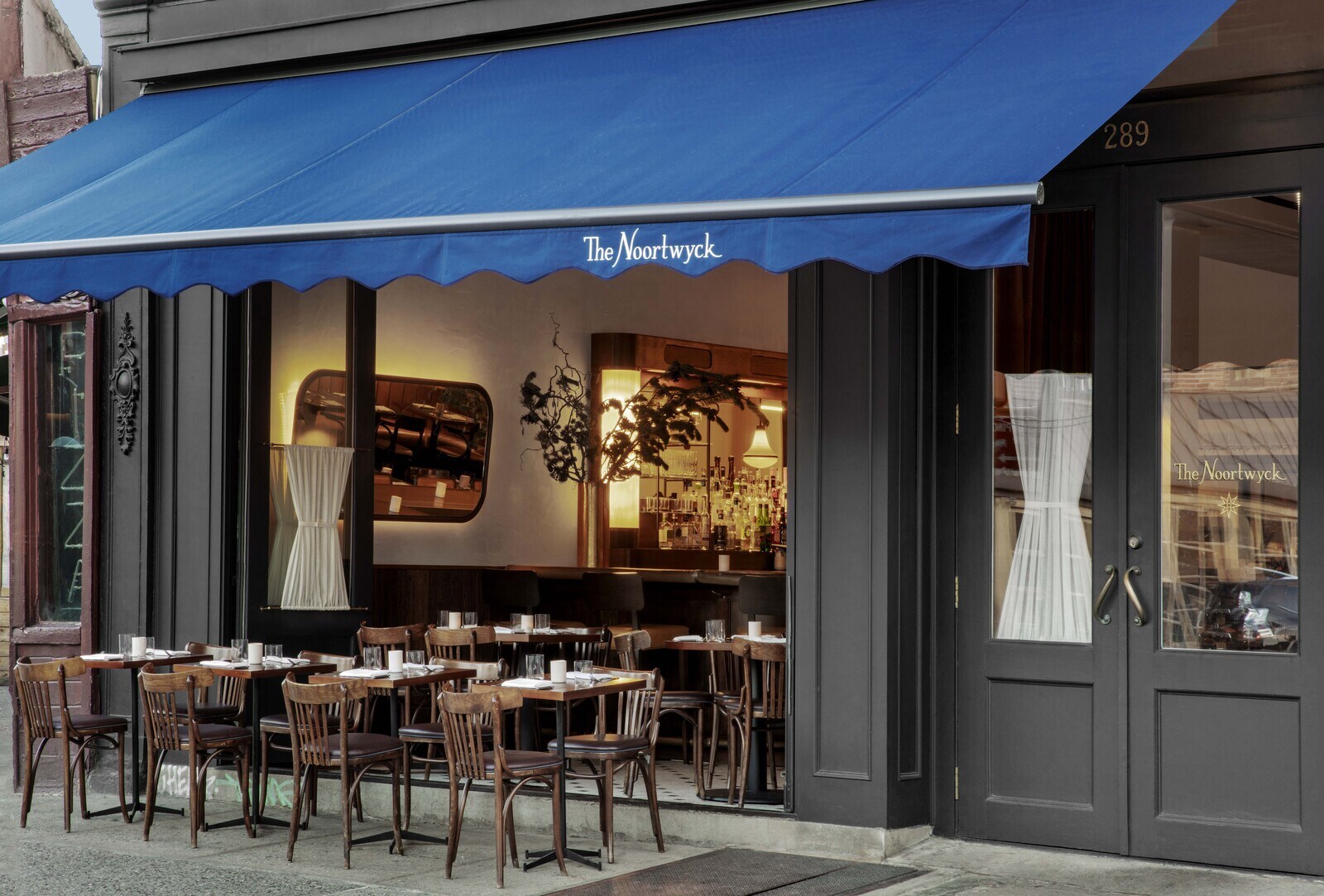 Glass doors in front of The Noortwyck open so that small tables and chairs are arranged both inside and outside under a blue awning. 