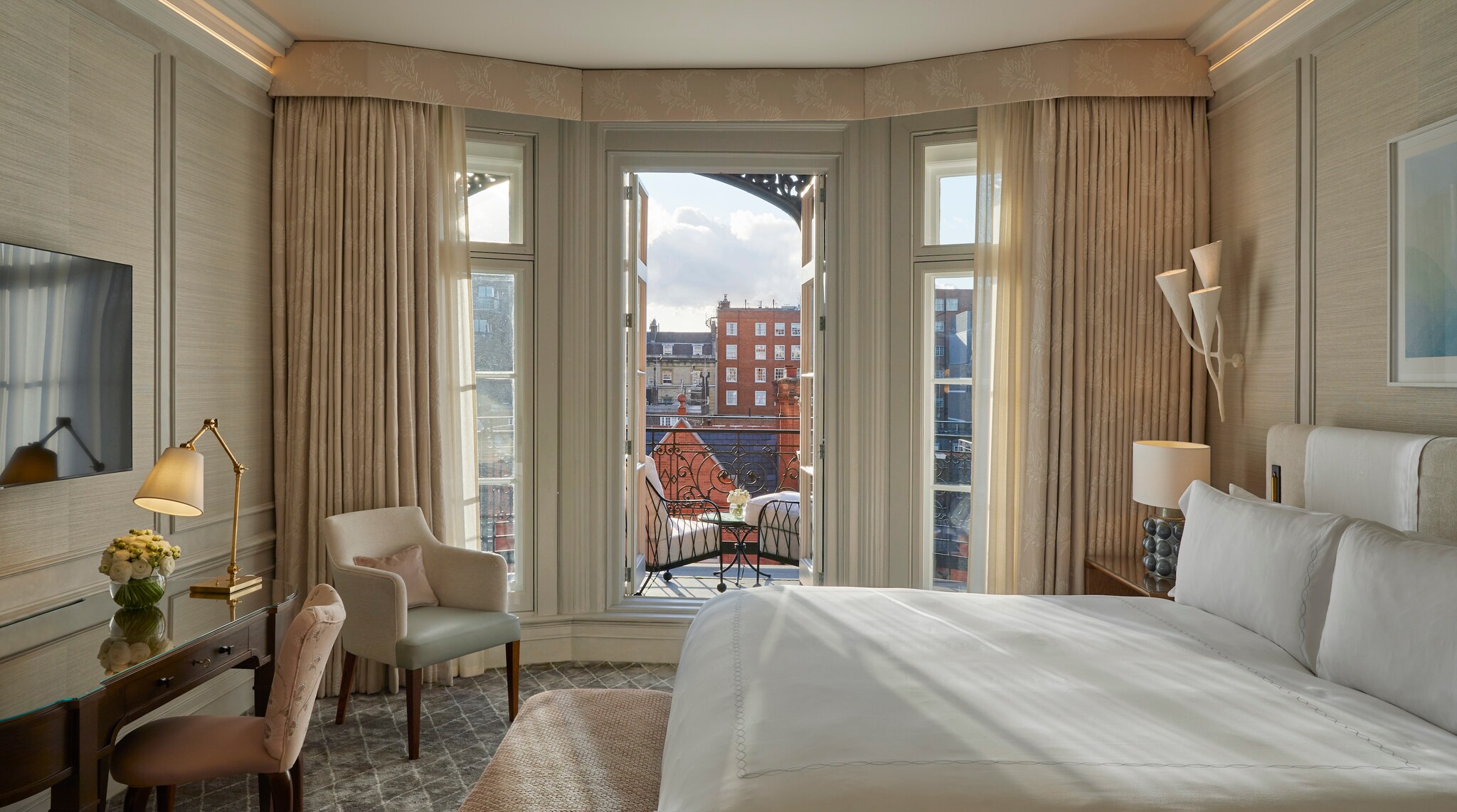 A room is decorated in white, cream, and gray tones. A large bed faces a desk and wall-mounted TV. Glass doors open onto a small balcony. 