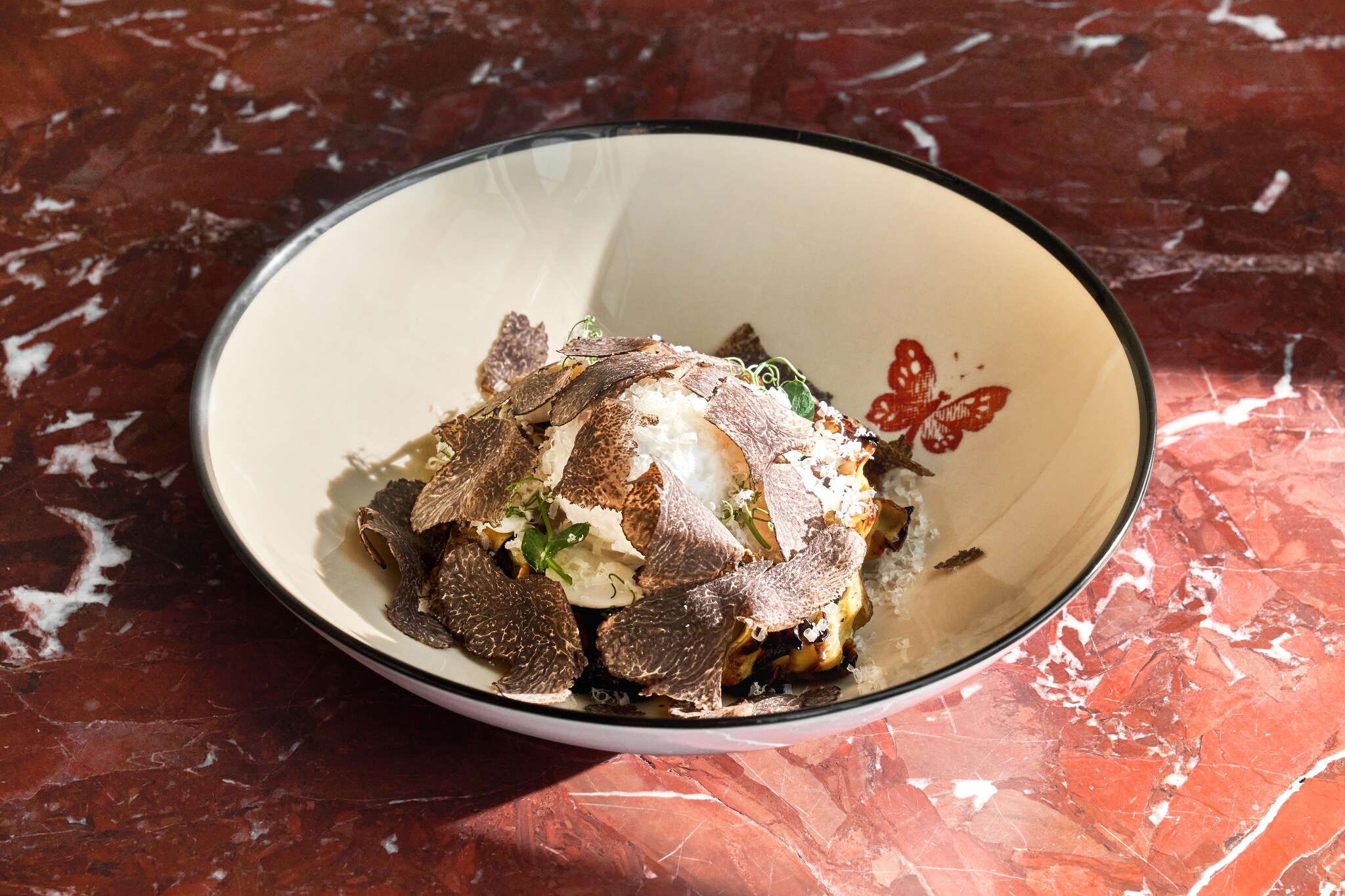 A white dish on a red marble table is filled with white cheese, herbs, and shaved curls of black truffle.