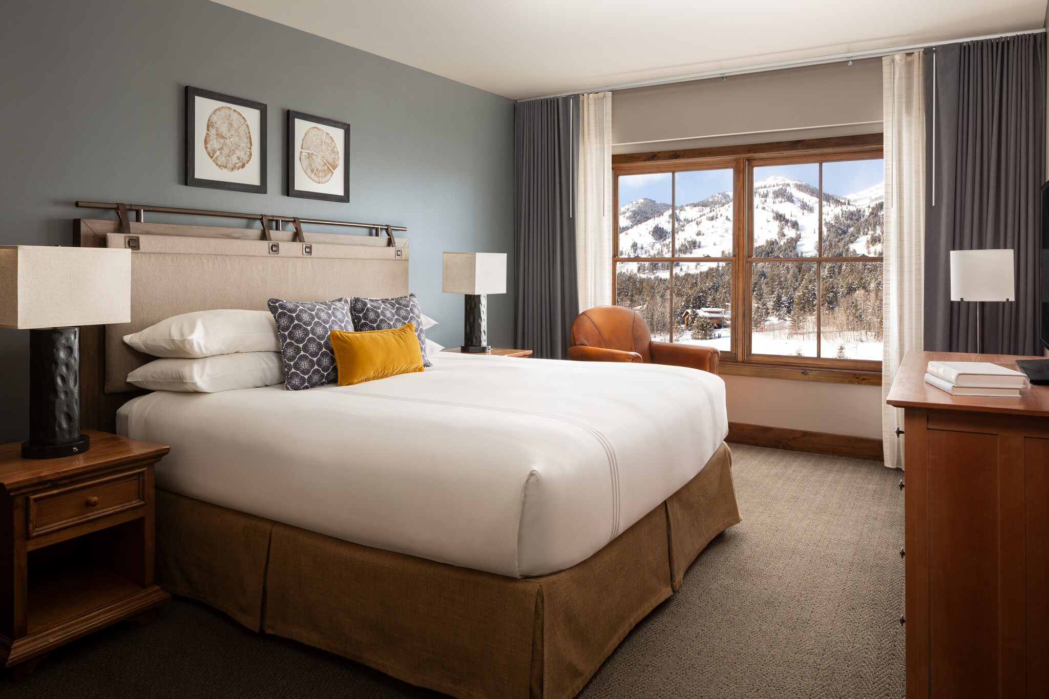 A guest room has a large bed with brown skirts, set against a dull green-gray wall. Wood-framed windows look out on a snowy mountain.