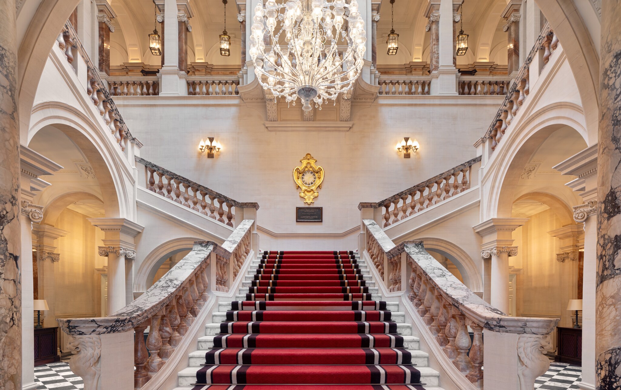 From the ground floor, the grand marble staircase climbs, then splits into left and right wings. It’s furnished with a red carpet.