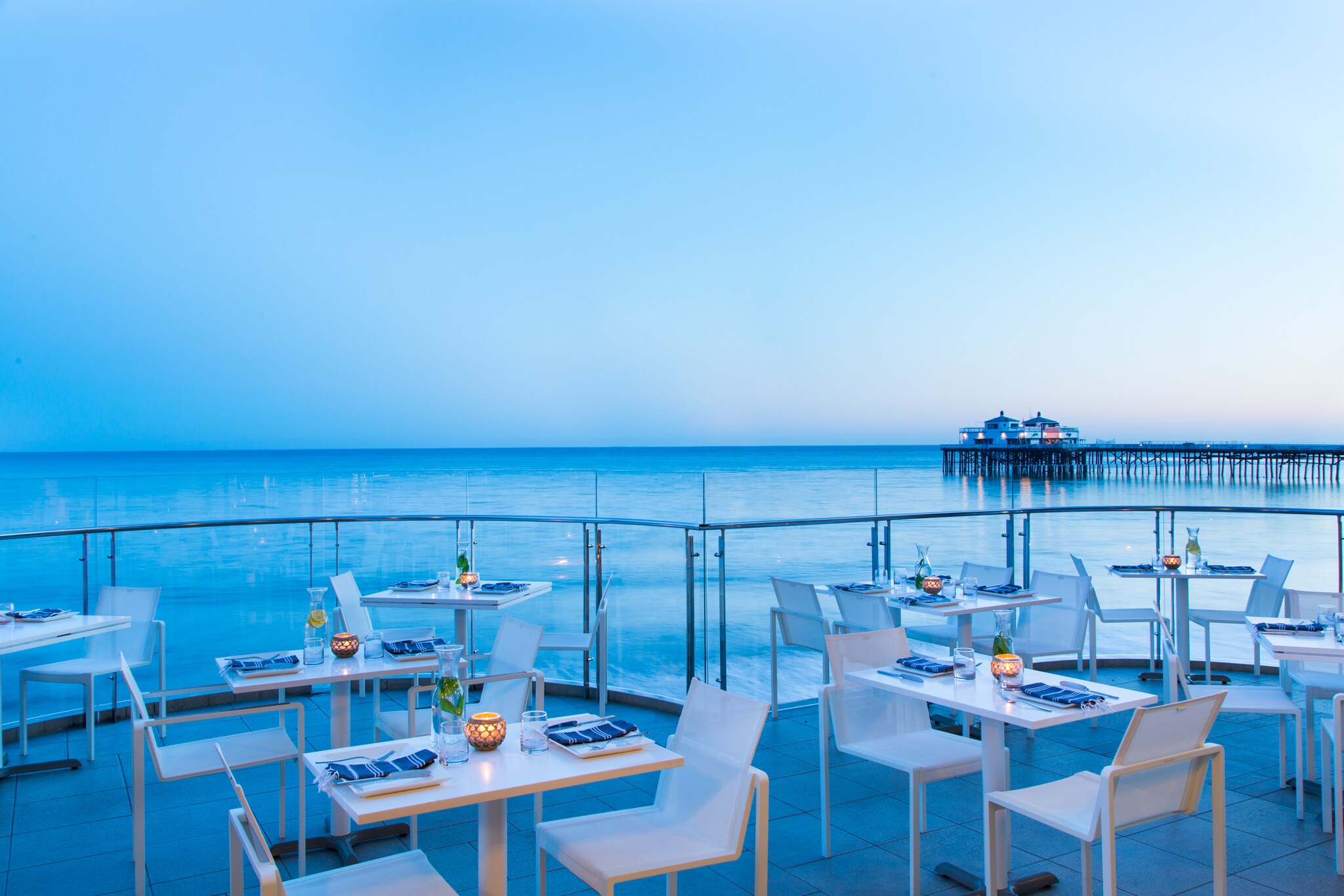 On a waterfront restaurant patio at Malibu Beach Inn, white tables and chairs are arrayed with small candles beneath a gloaming blue sky.