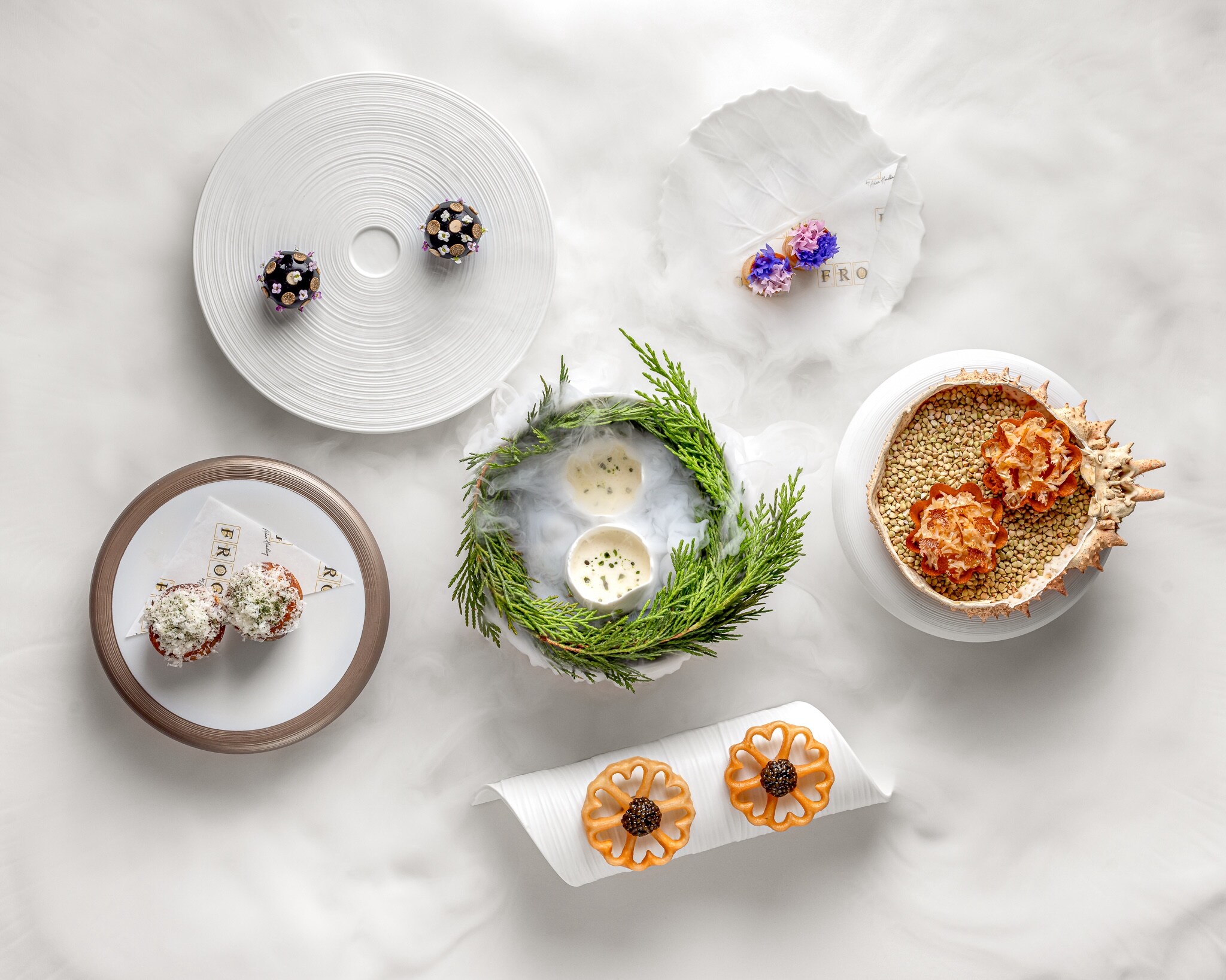 Overhead view of six dishes on a white marble tabletop. Most of the food consists of duos of small round pieces in various colors.