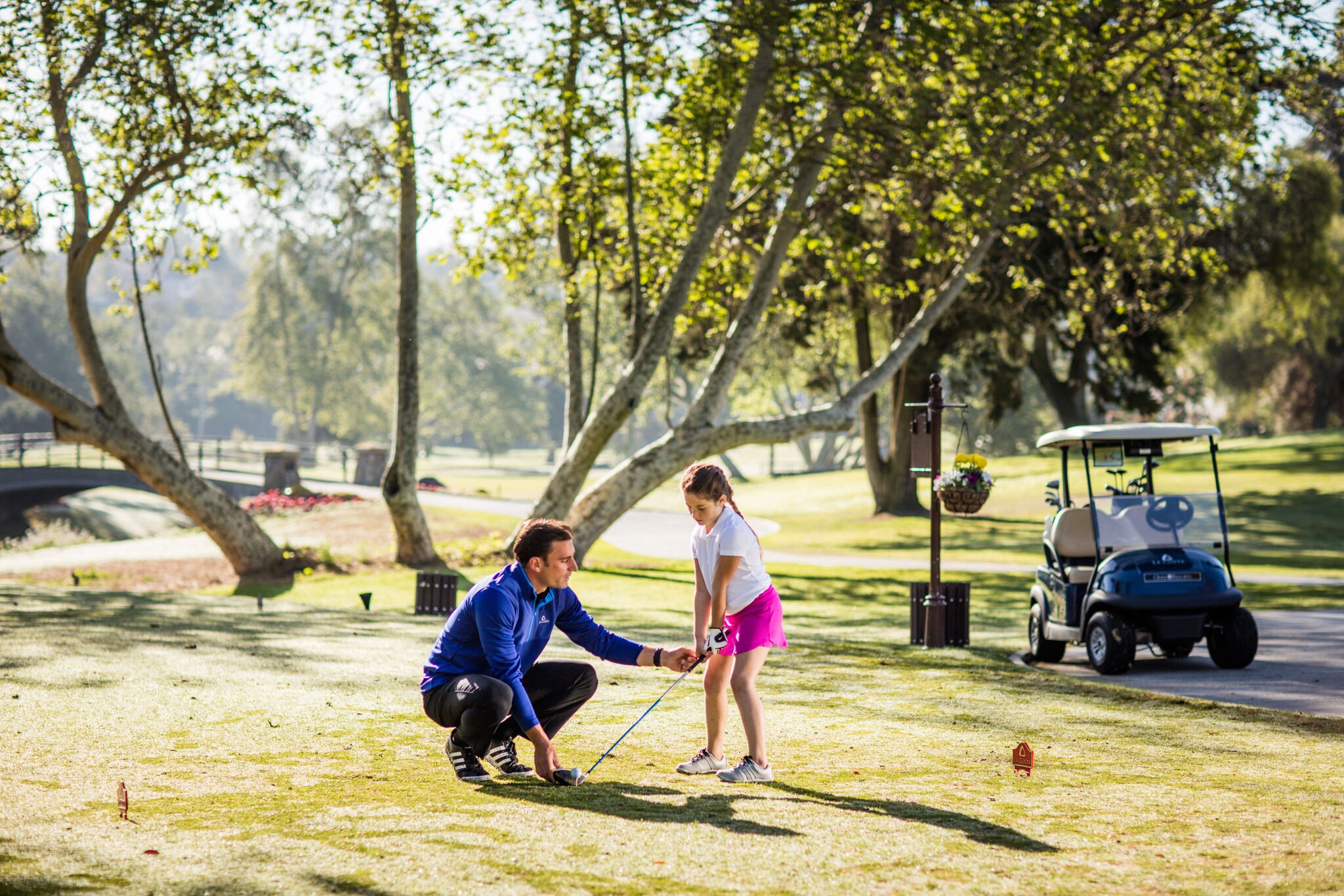 On a golf course, a white man crouches down to guide a young white girl as she positions her club behind the tee.