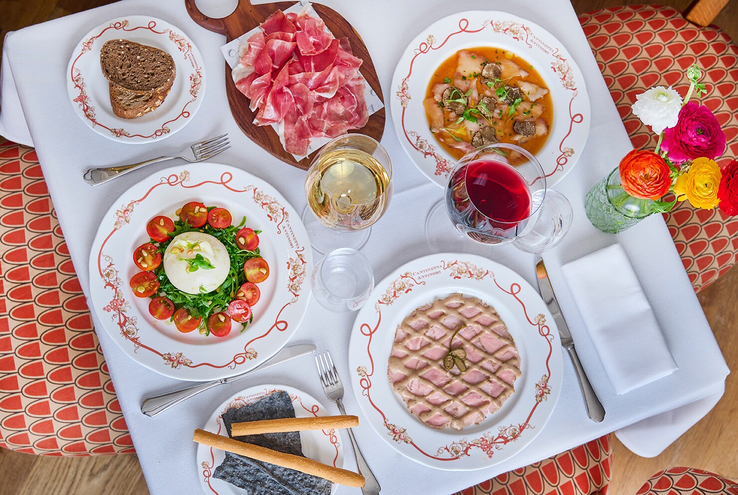 Overhead view of a white table covered with several dishes, including salad, soup, a pink waffle-like dessert, and a board of prosciutto.