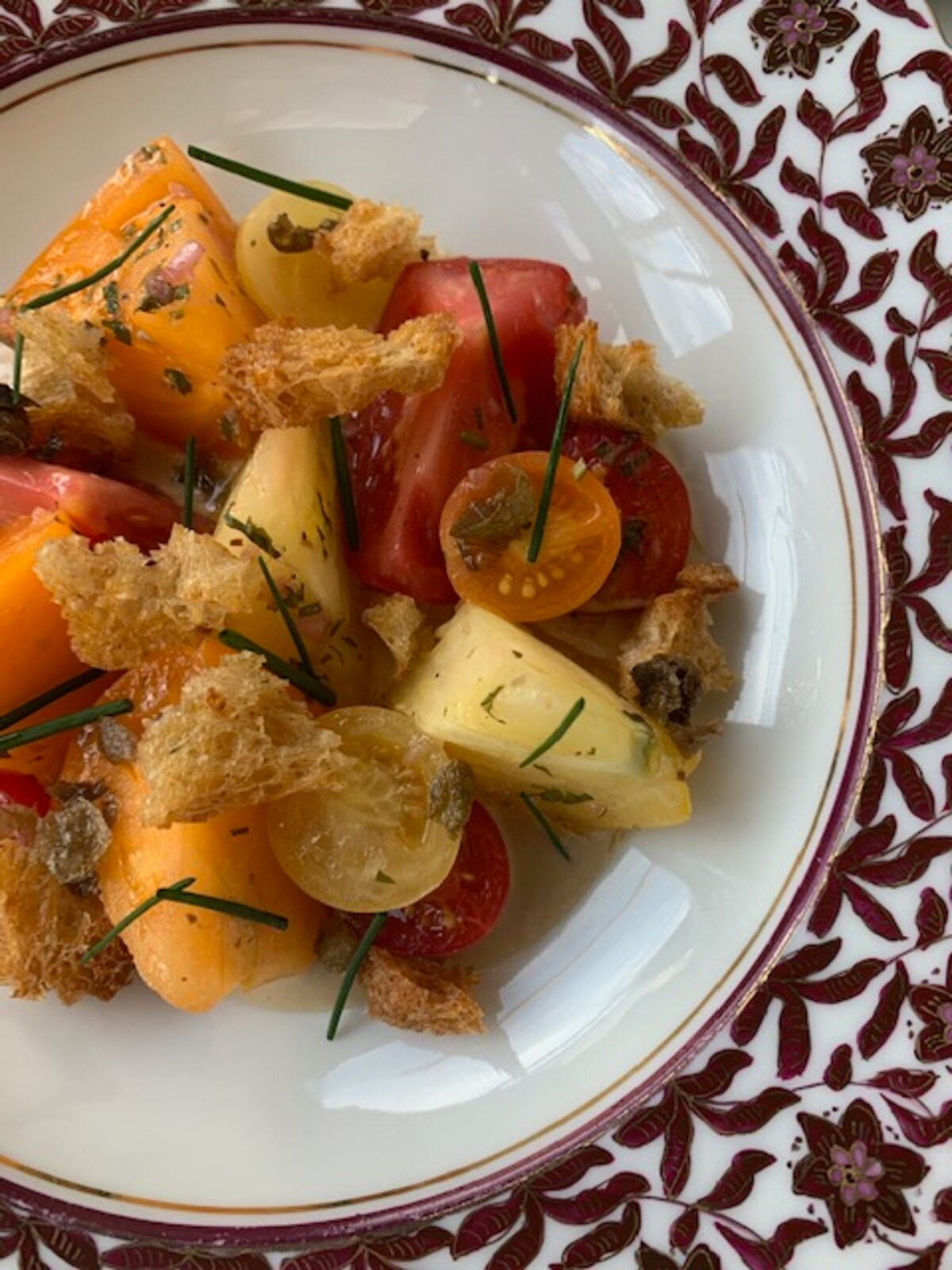 On a white dish with dark red floral trim, thick wedges of tomatoes are tossed together with capers, tonnato sauce, and airy croutons.