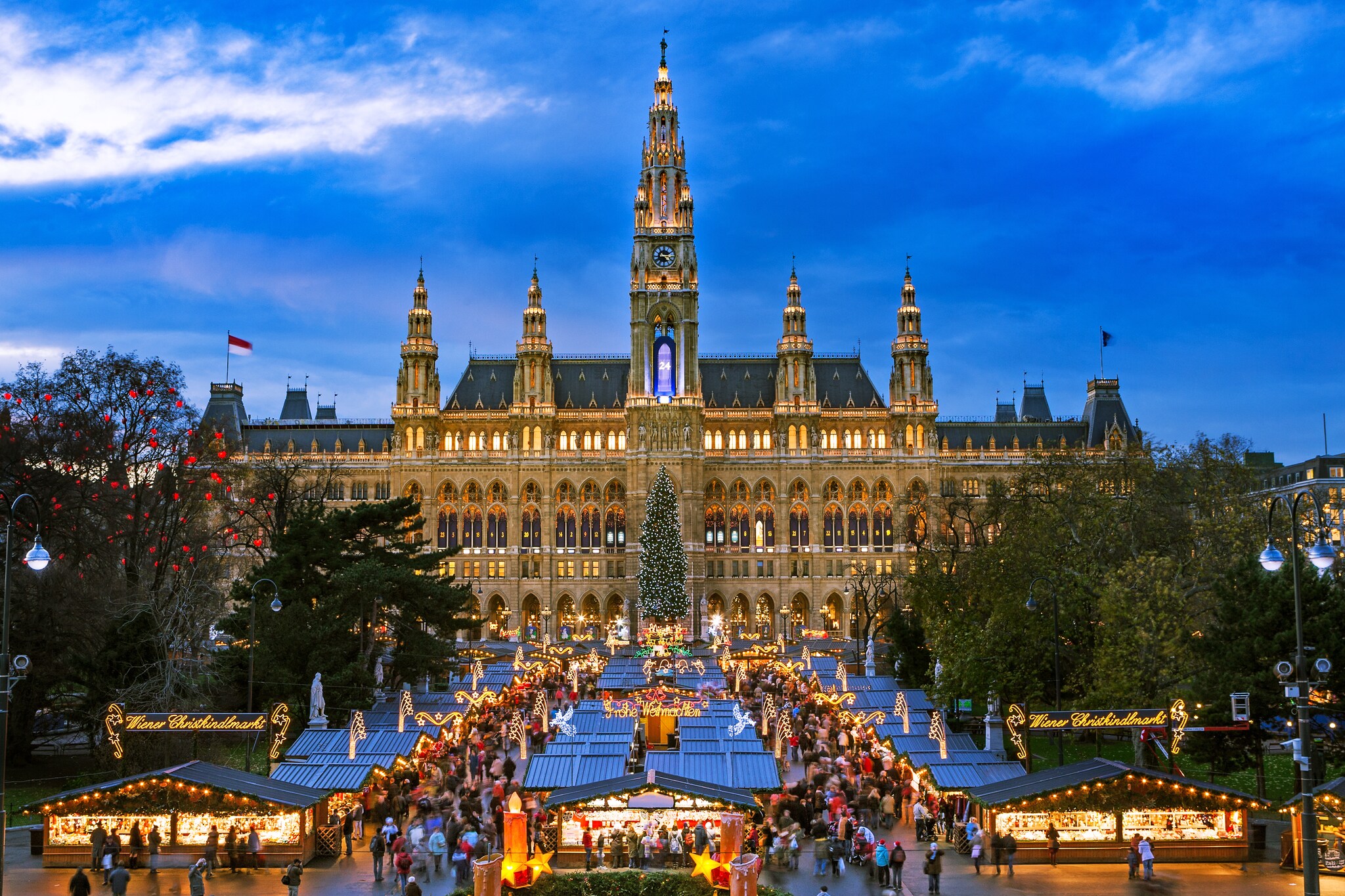 In early evening. Vienna City Hall looms over rows of small pop-up sheds strung with lights and crowded with shoppers at a Christmas market.