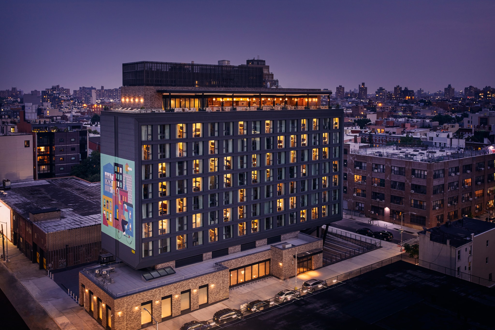 Exterior of the hotel in the evening. Against a purple sky over Williamsburg, windows in the boxy, modern hotel glow golden.
