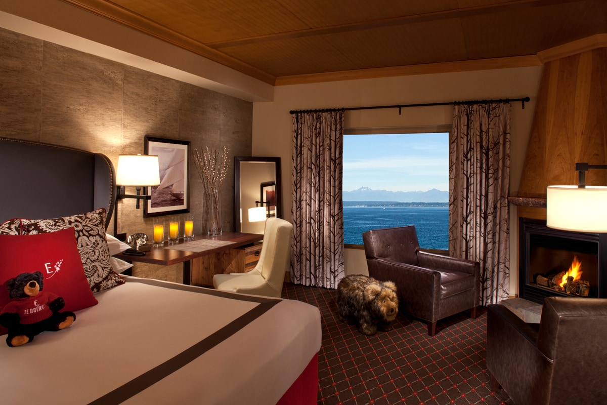 A Waterside Guestroom has earth-toned ceiling and walls, a teddy bear on the bed, a fireplace, and a window overlooking the Puget Sound.
