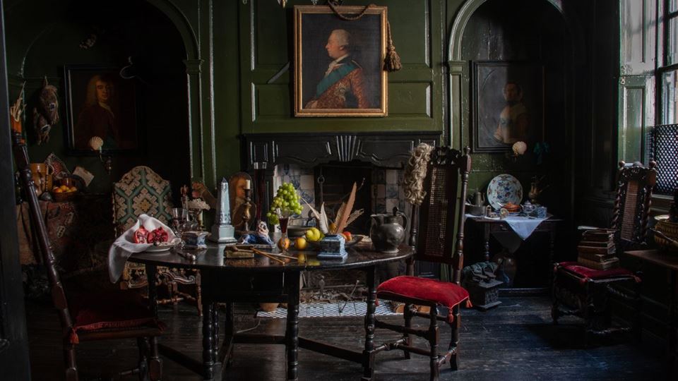The living room of Dennis Severs’ House is dim, with olive-green walls, a portrait over a black fireplace, and a table piled with fruits.